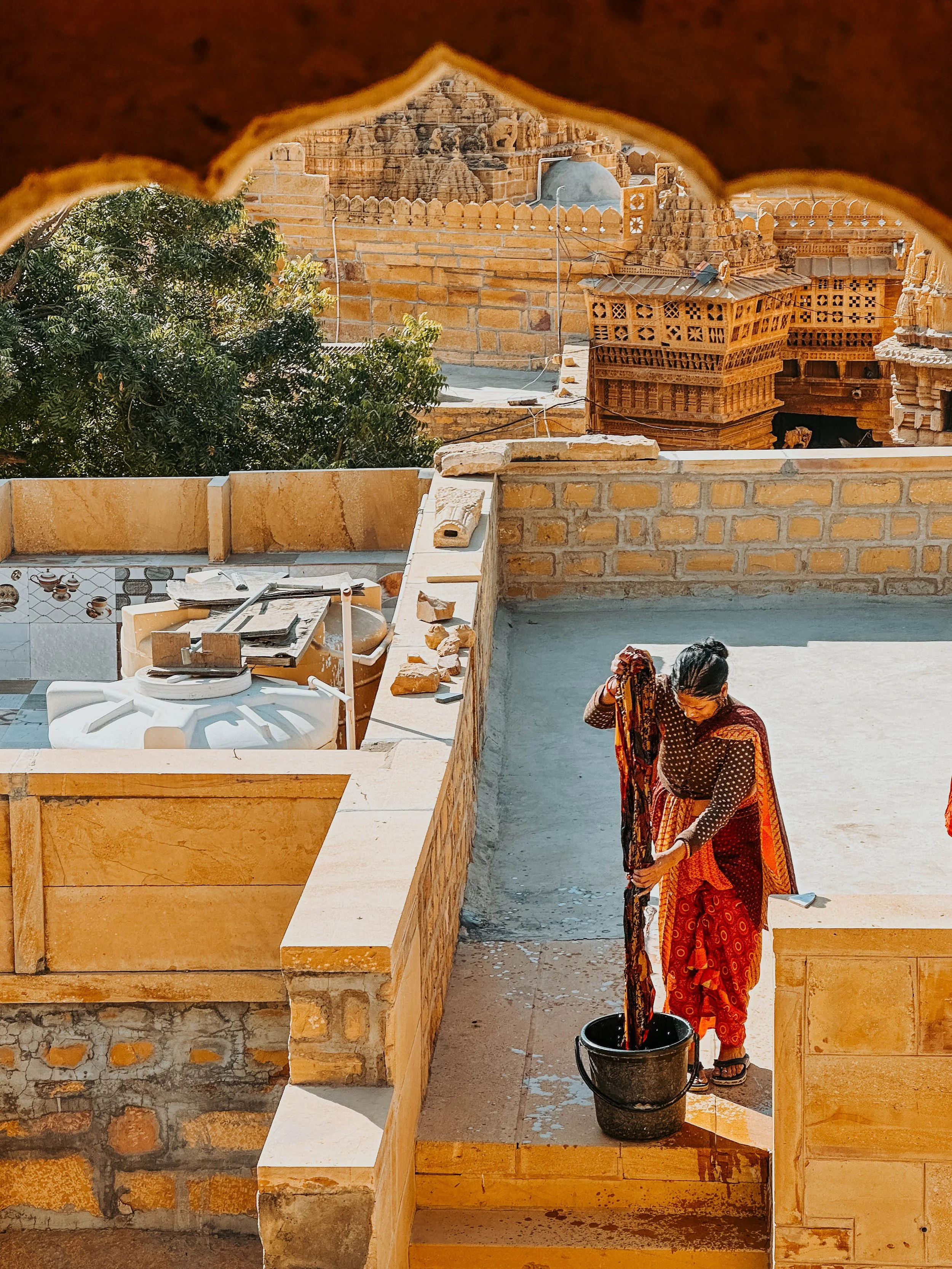Una donna indossa un sari colorato mentre lavora i tessuti su un terrazzo di un edificio storico in India, con un muro di mattoni e strutture architettoniche in stile tradizionale.