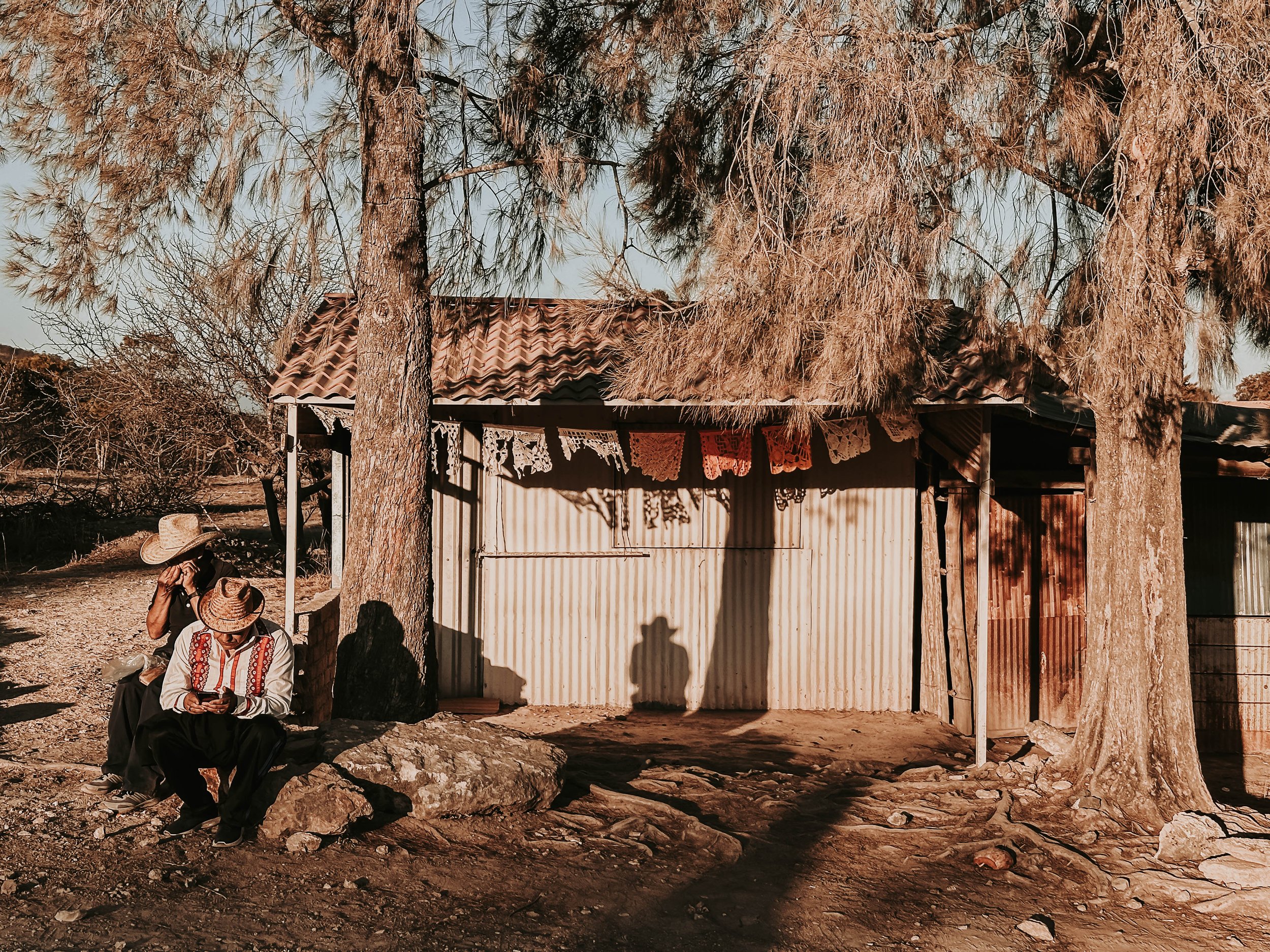 Due persone sedute su una roccia, indossano cappelli di paglia e abiti tradizionali, all'aperto vicino a una capanna in legno con tetto di tegole, tra alberi, in un ambiente rurale