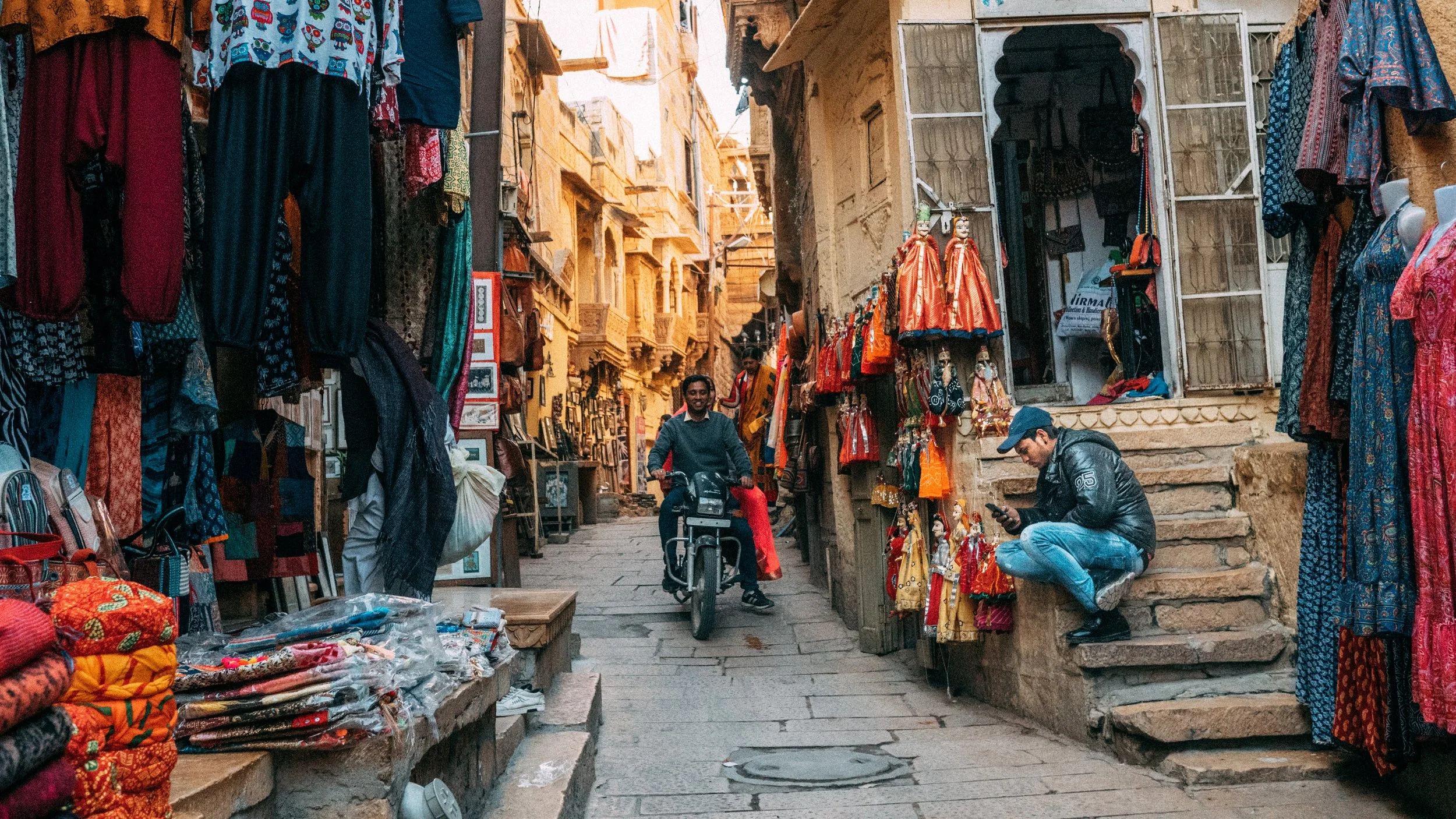 Strada stretta in un mercato con bancarelle di tessuti e abbigliamento colorato, uomo in bicicletta e uomo seduto sui gradini che guarda il cellulare.