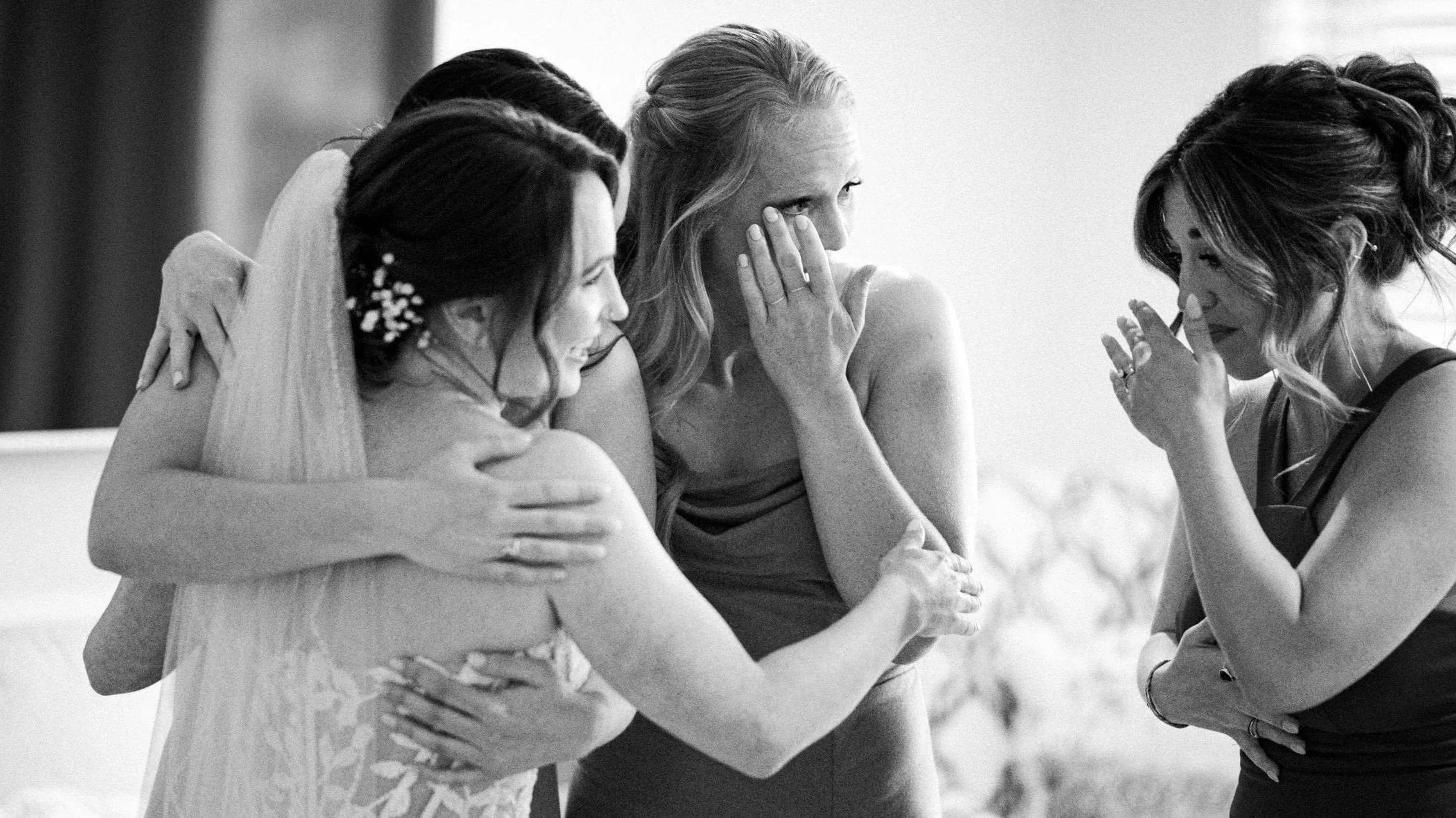 a candid moment of a bride with her bridesmaids group hugging and wiping happy tears with the first look of the brides dress