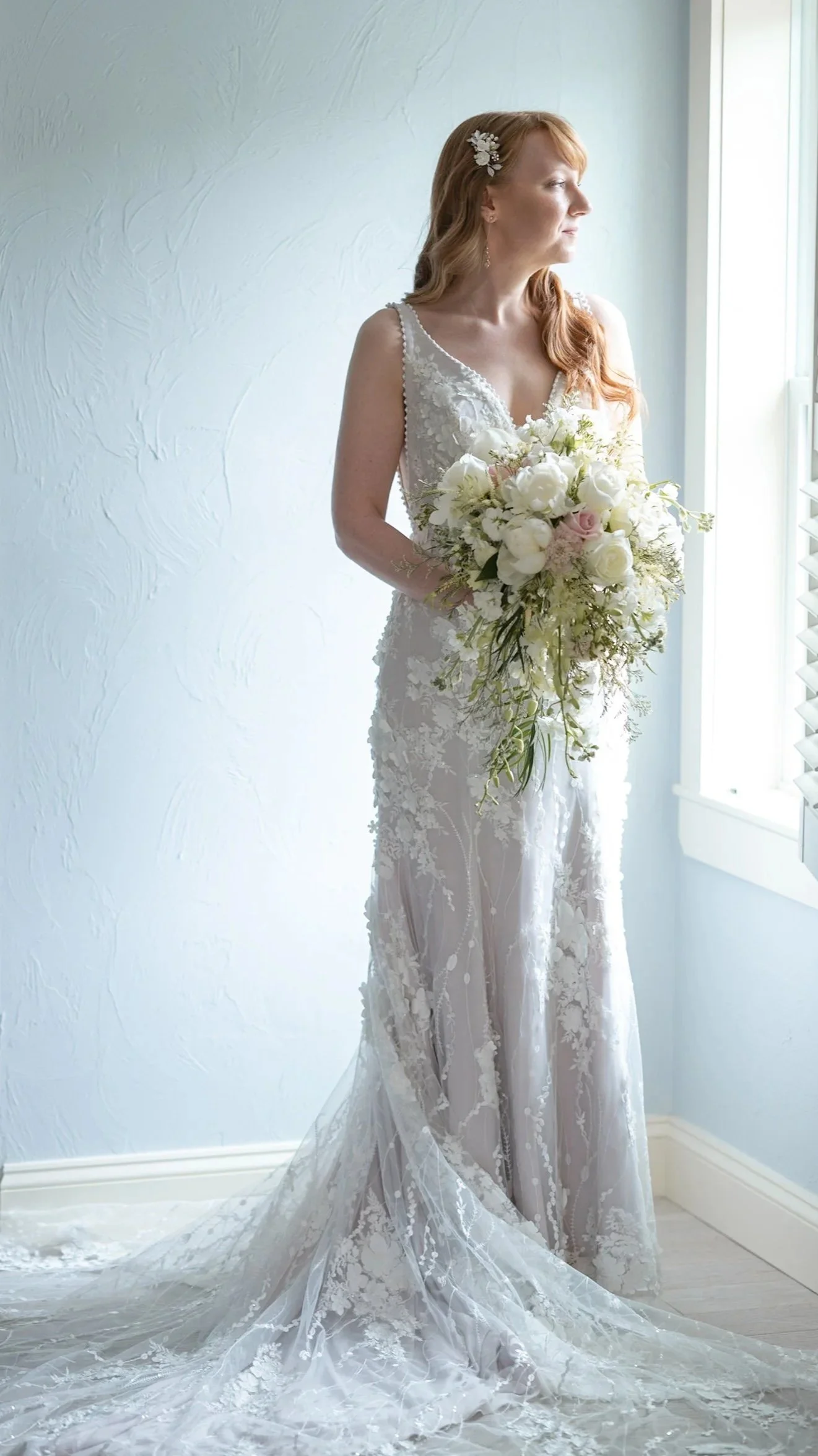 Bride in a white lace wedding dress, holding her bouquet looking out a window