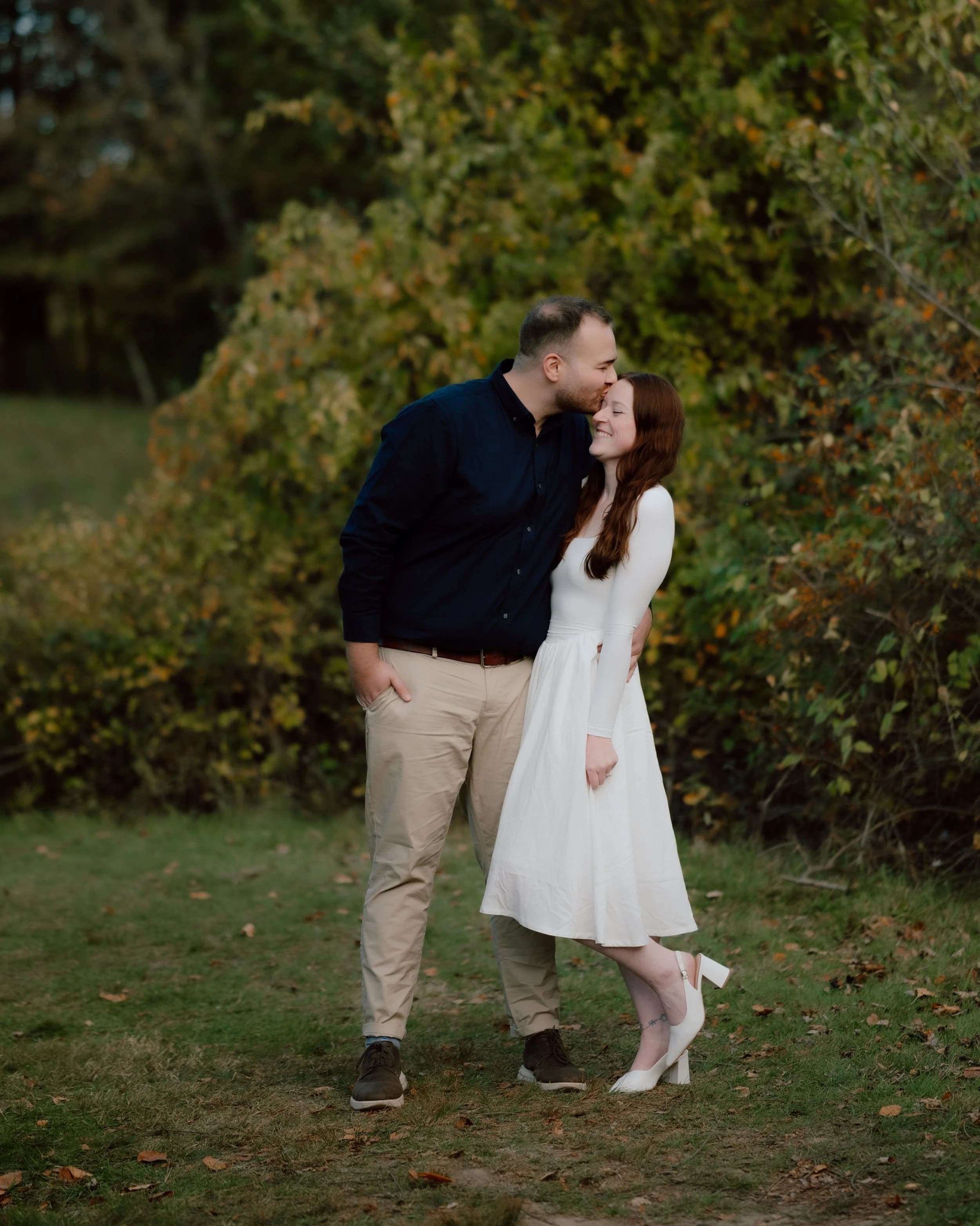 A man and woman standing close together on a grassy area with trees and bushes in the background, smiling and leaning toward each other, with the man's forehead touching the woman's forehead.