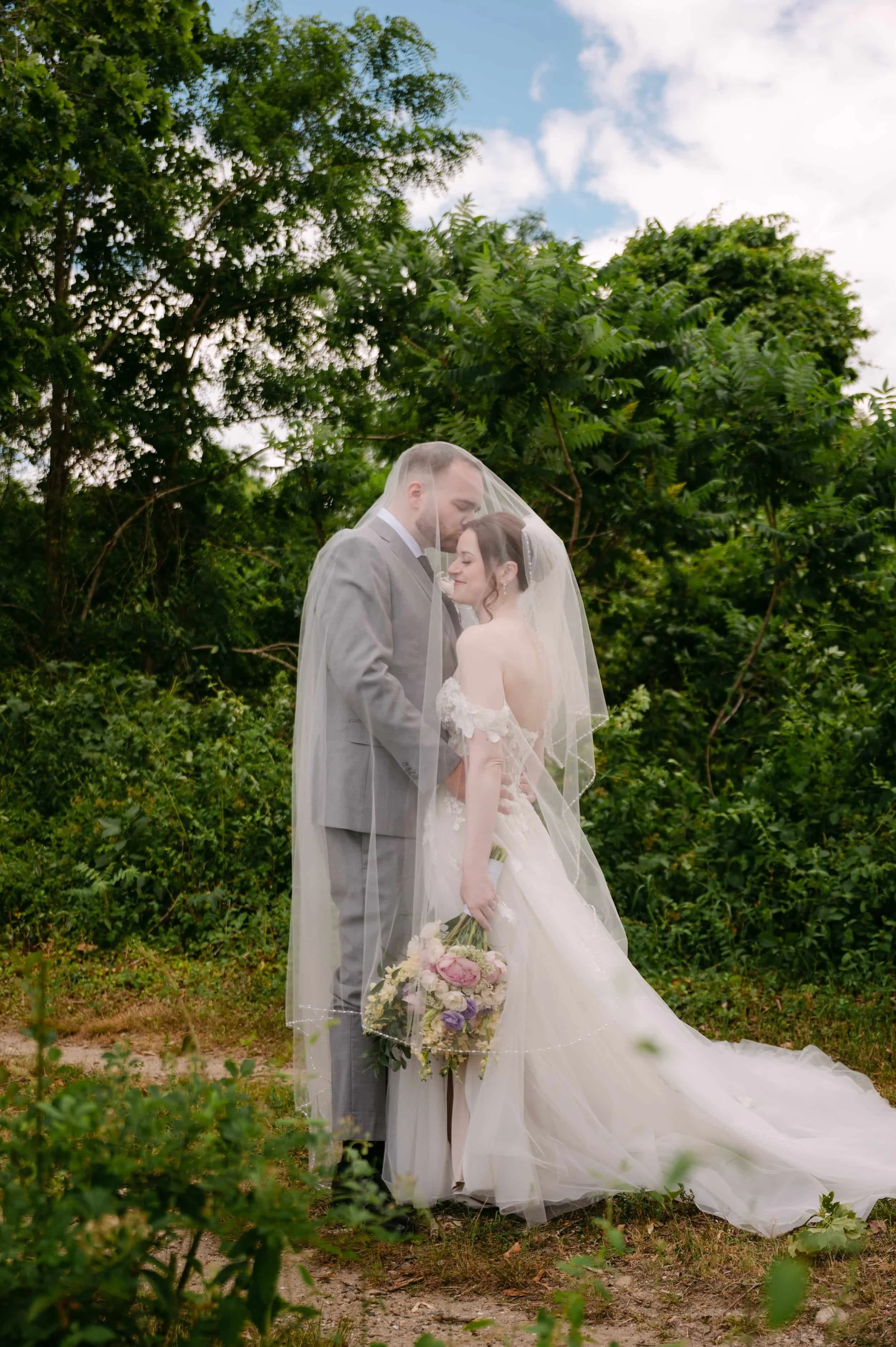 A bride and groom standing close together outdoors, sharing a tender moment under a veil, with lush green trees and a blue sky in the background. The bride has dark hair, and the groom has short hair and is wearing a light gray suit. The bride is holding a bouquet of flowers.
