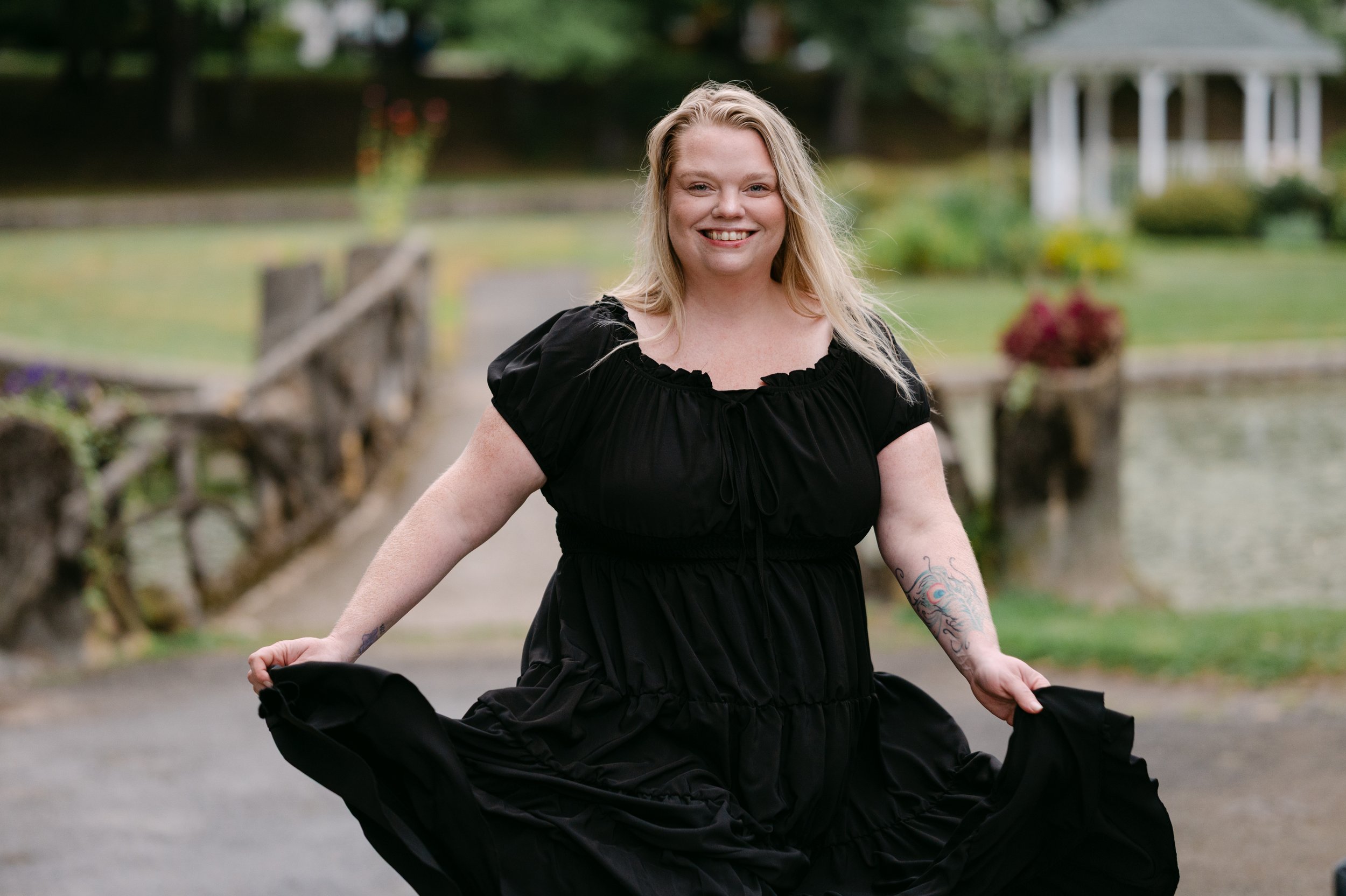 A smiling woman with long blonde hair, tattoos on her arms, wearing a black dress, standing outdoors on a path near a body of water with a gazebo in the background.