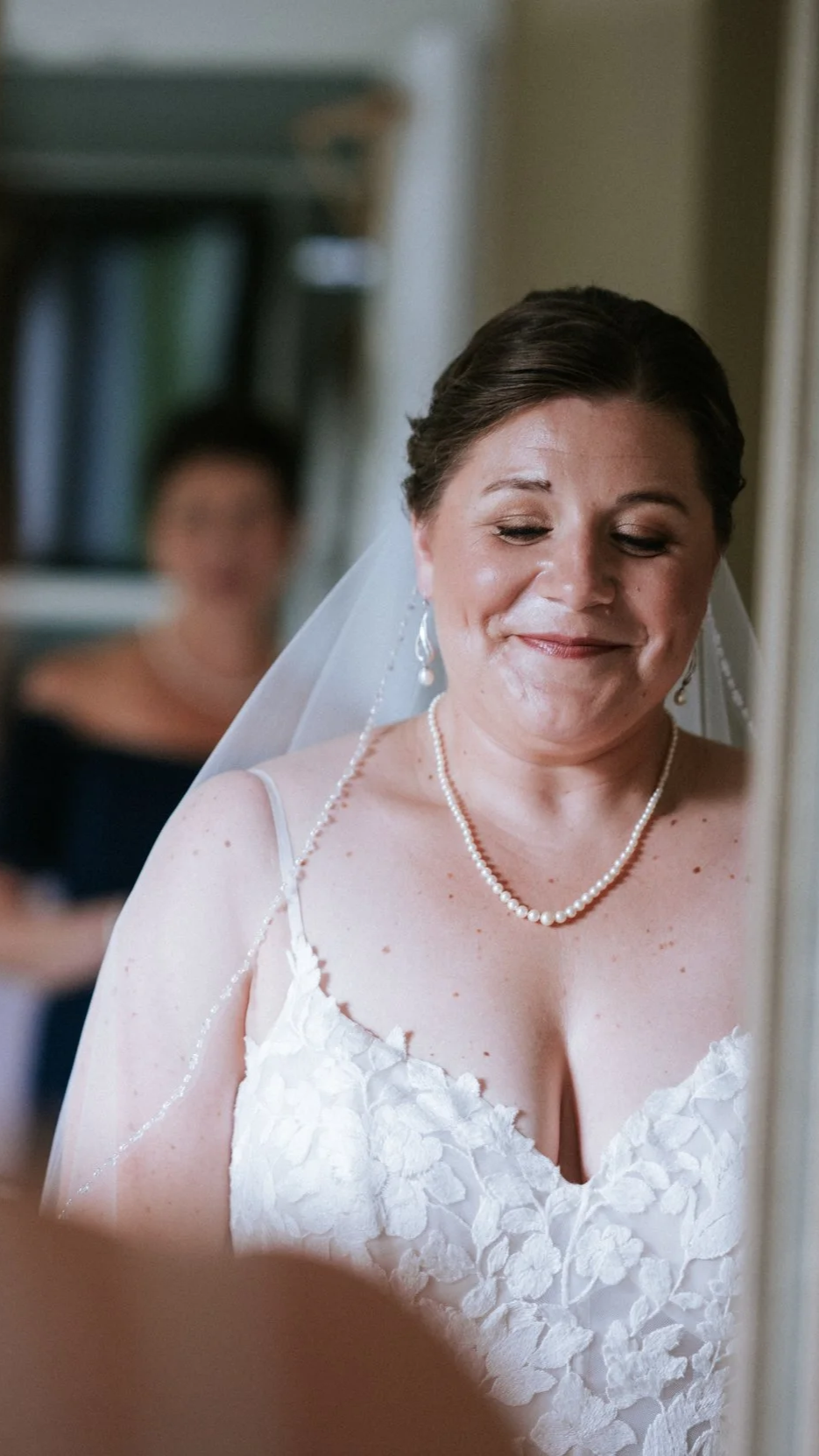 A bride with short dark hair, wearing a white lace wedding dress, pearl necklace, earrings, and veil, smiling softly as she looks down.