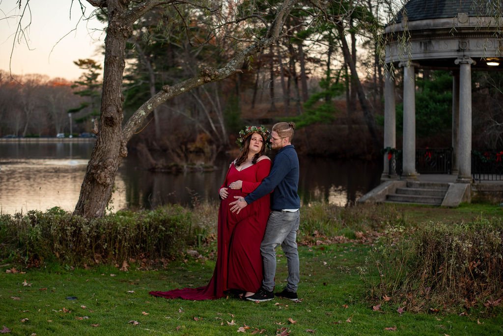 A couple standing near the pond at Slater memorial park in Pawtucket, RI during sunset, with the woman wearing a red gown and floral crown, and the man in a blue jacket and gray pants, embracing
