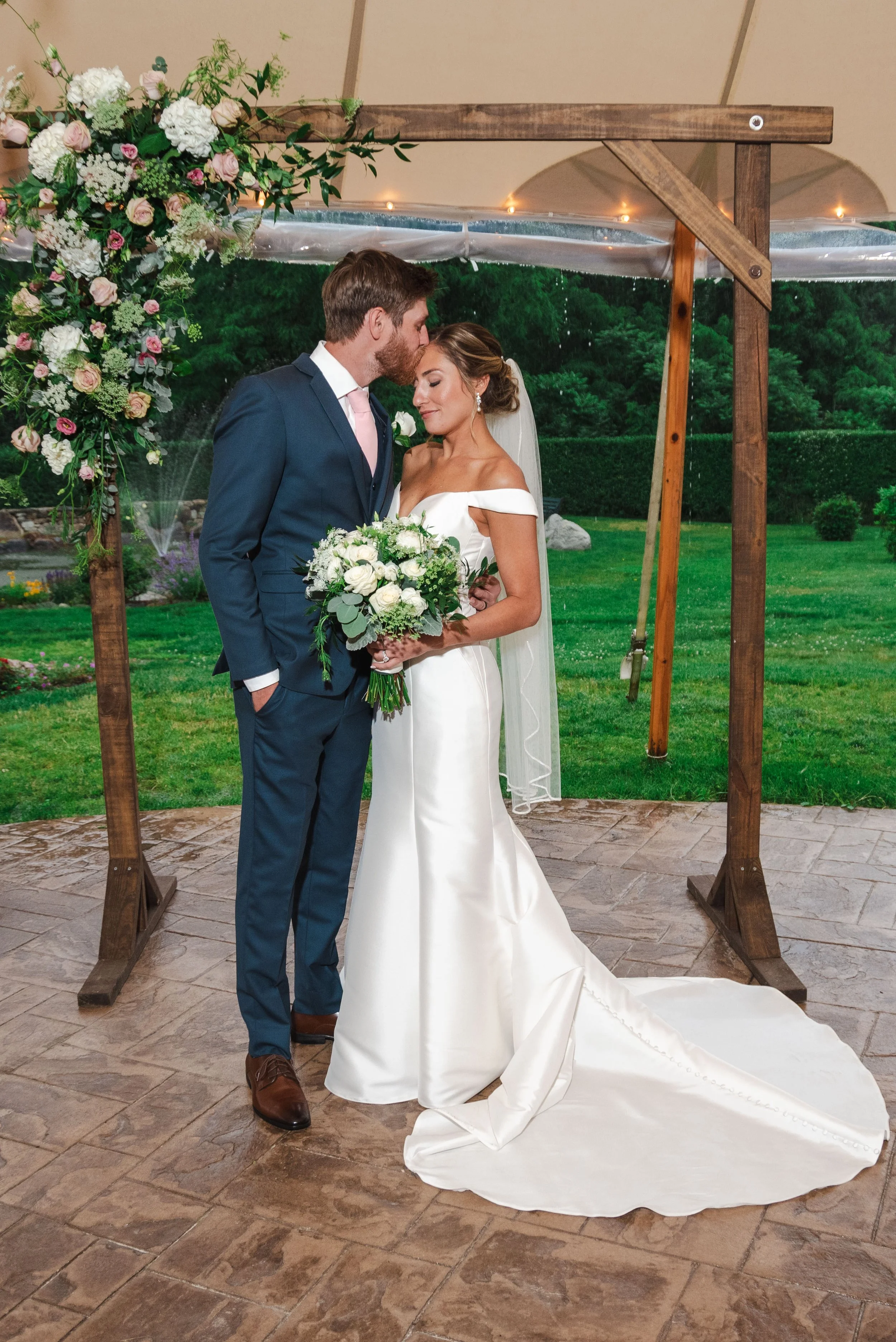 A bride and groom standing close together under a wooden arch, with the groom kissing the bride on the forehead. The bride is wearing a white wedding gown and holding a bouquet of white and green flowers. The groom is dressed in a dark blue suit with a light pink tie. The scene is outdoors with a lush green background.