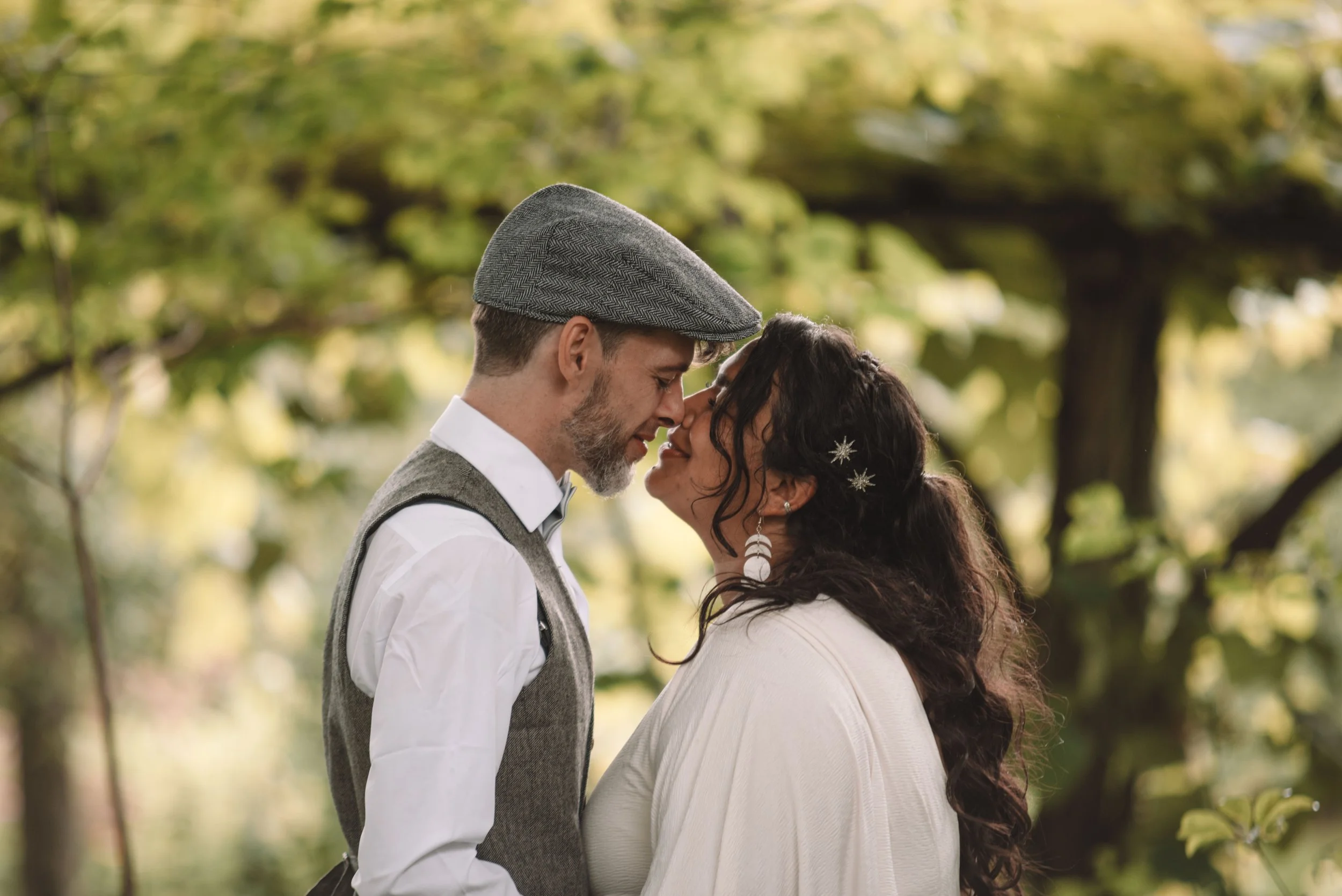 A couple about to kiss outdoors in a park with green trees in the background.