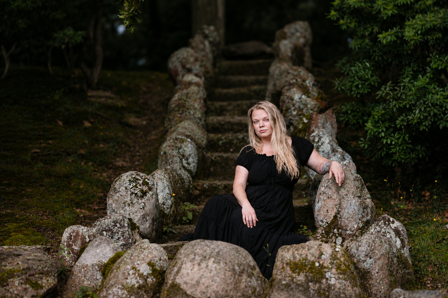 A woman with long blonde hair sitting on stone stairs outdoors, surrounded by moss-covered rocks and greenery.