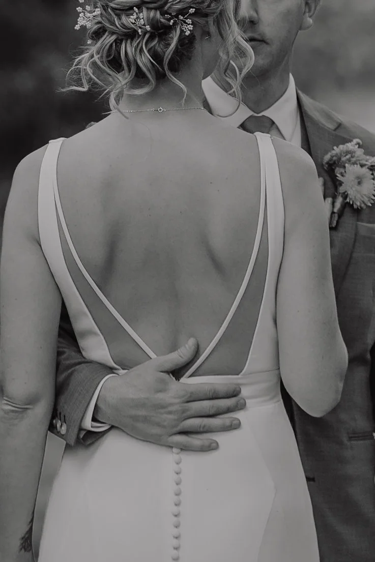A bride and groom embrace during their wedding, with the bride's back visible showing her elegant dress and the groom’s hand resting on her waist.