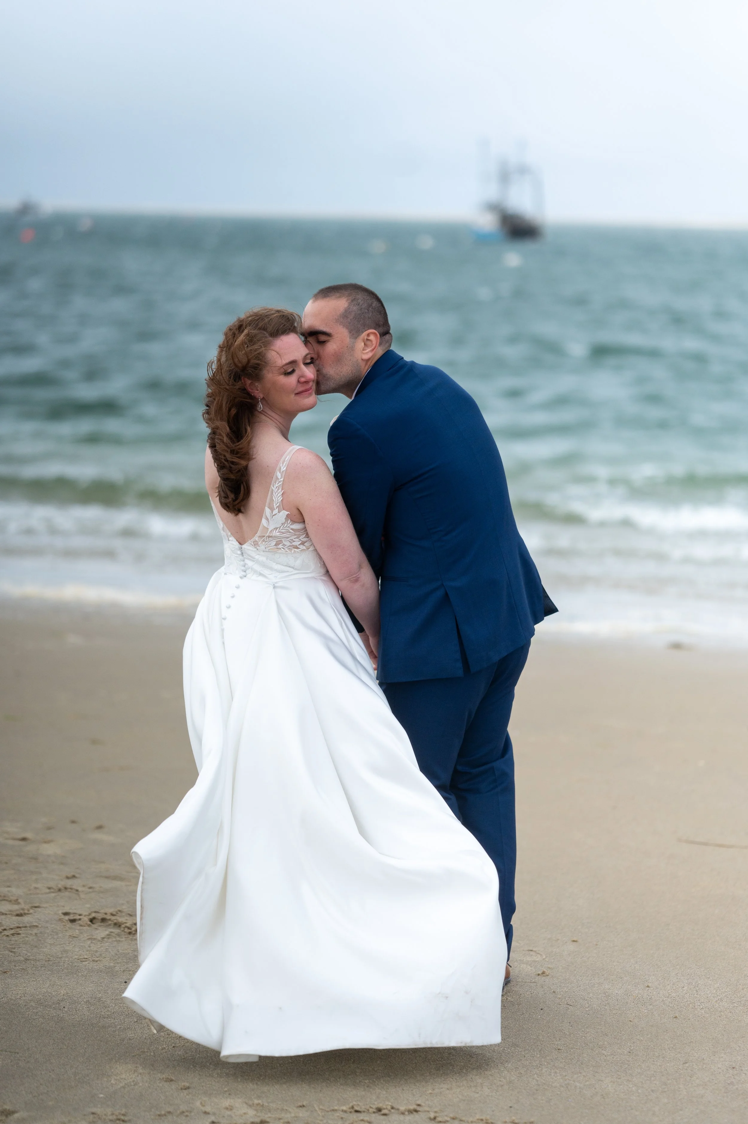 A bride and groom sharing an intimate moment on the beach, with the groom kissing the bride's forehead as they stand close to the shoreline.