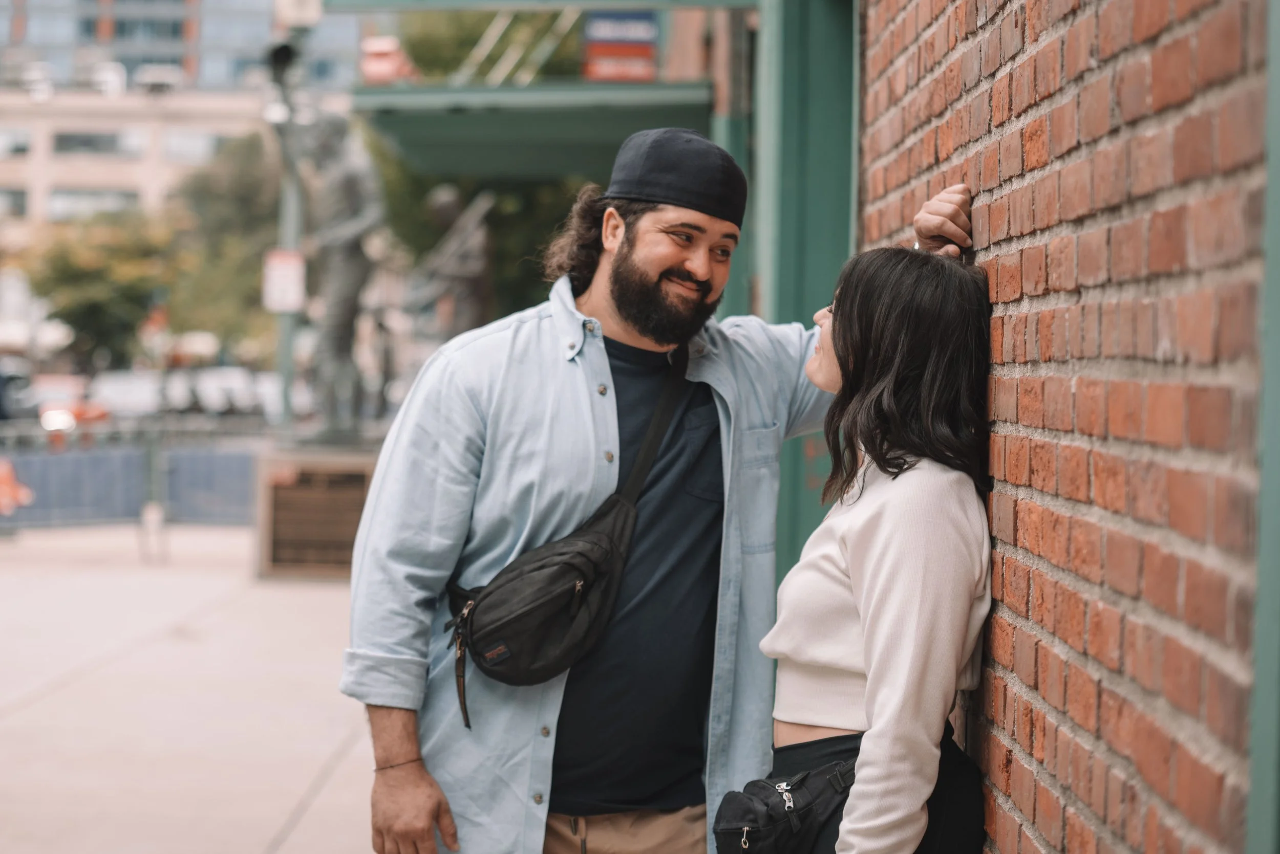 A newly engaged couple are standing close to each other outside of Fenway park in Boston, with the man leaning against a brick wall and smiling at the woman. The woman is partly leaning against the wall, and they are looking into each other's eyes.