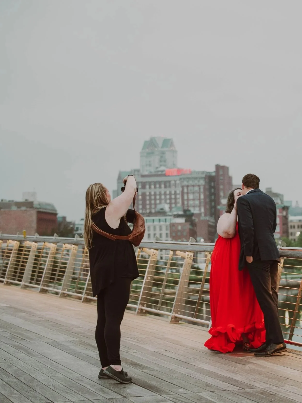 A woman in a red dress and a man in a dark suit are kissing on a rooftop with a cityscape in the background. A woman is taking a photo of them.