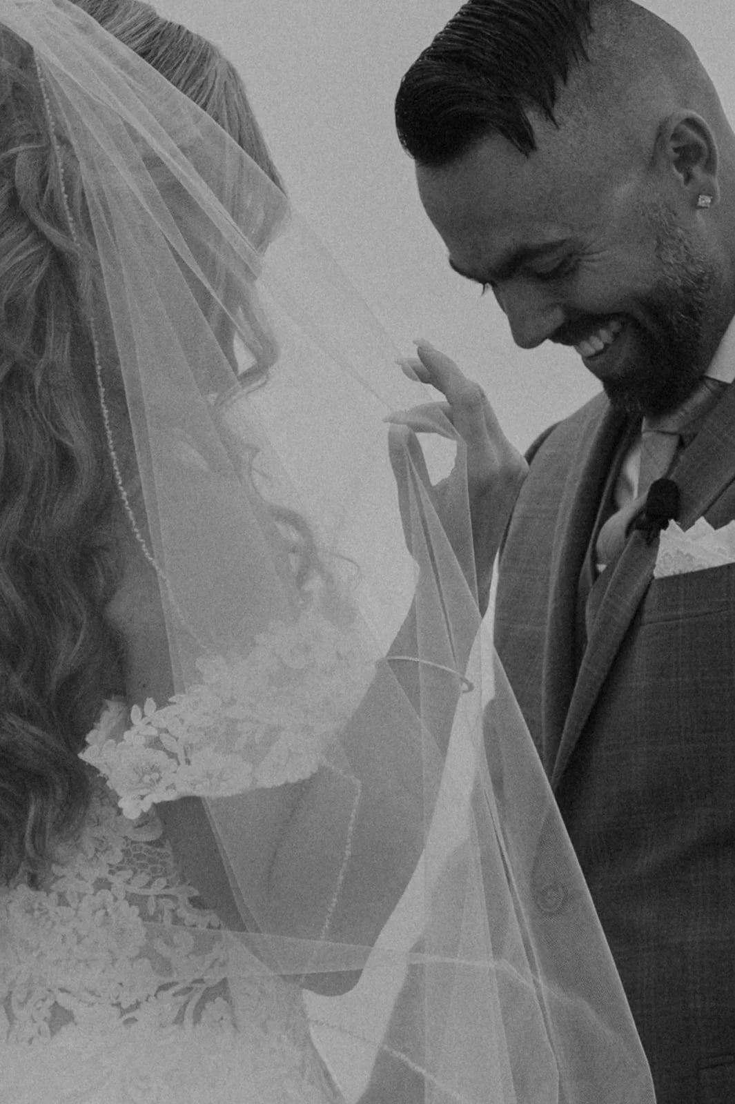 Black and white photo of a bride and groom smiling, with the bride holding a bouquet and the groom lifting her veil.