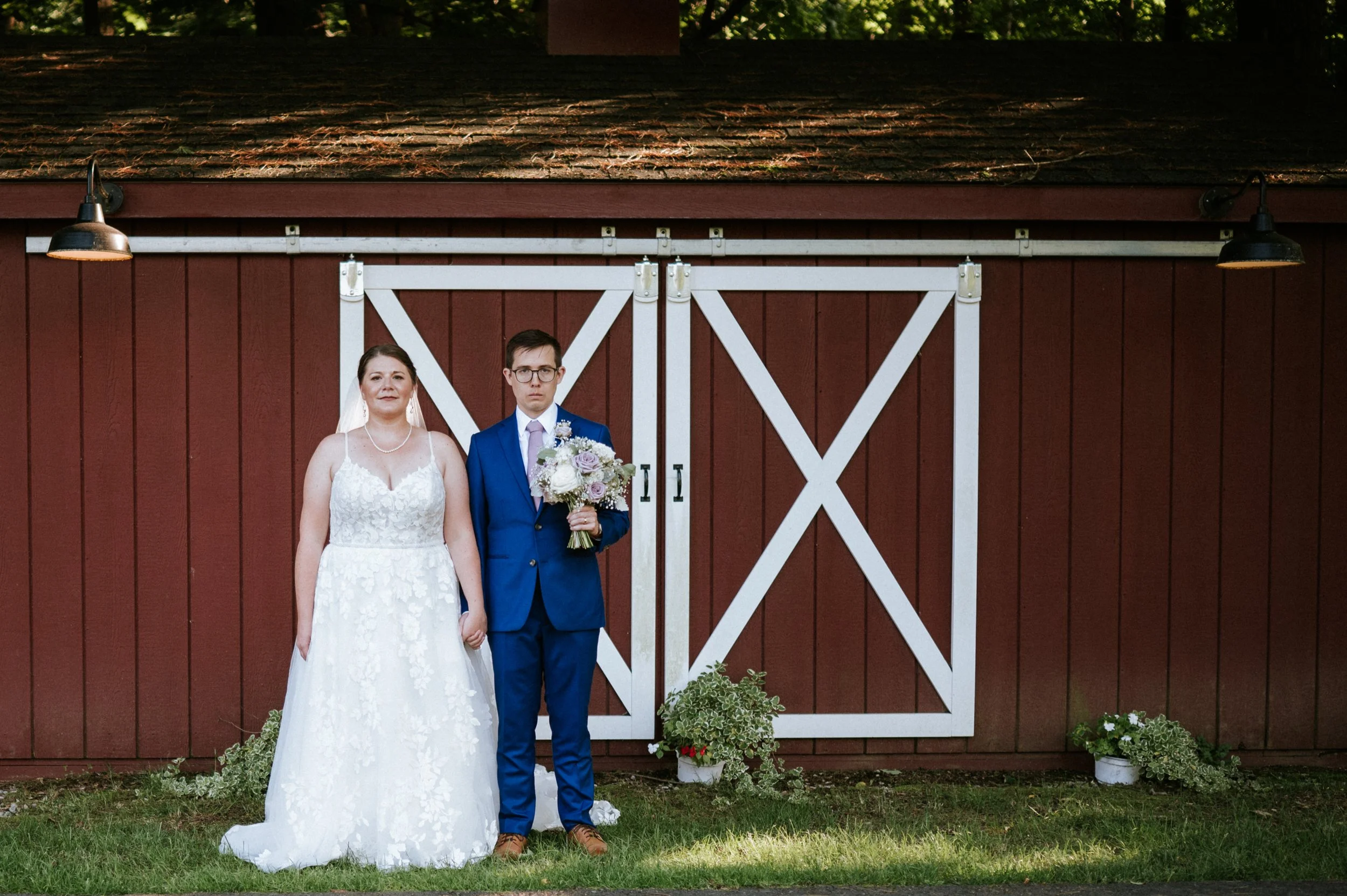 A bride and groom standing outside in front of a red barn with white doors, green grass, potted plants, two wall-mounted lights, and trees in the background.