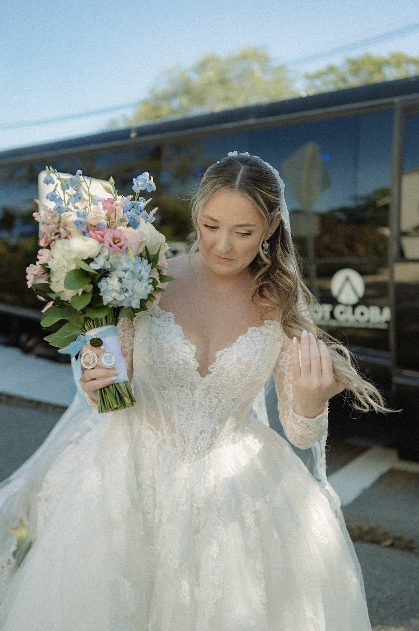 Bride in a lace wedding dress holding a bouquet, standing outdoors near a bus, looking down with a gentle smile.