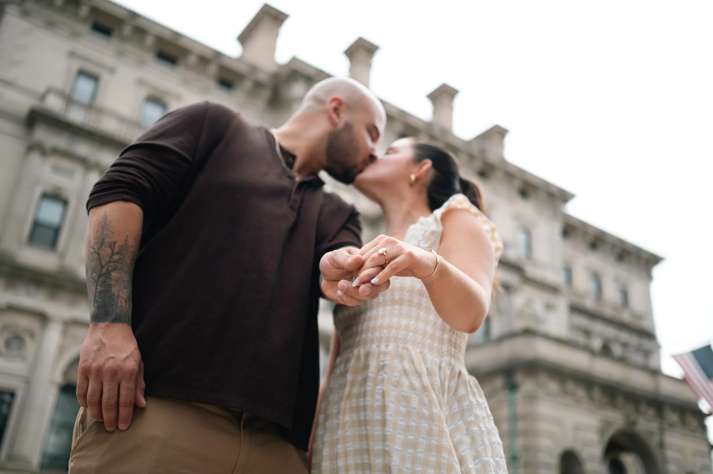 A couple kissing in front of The Breakers in Newport RI, holding hands, with the woman showing her engagement ring.