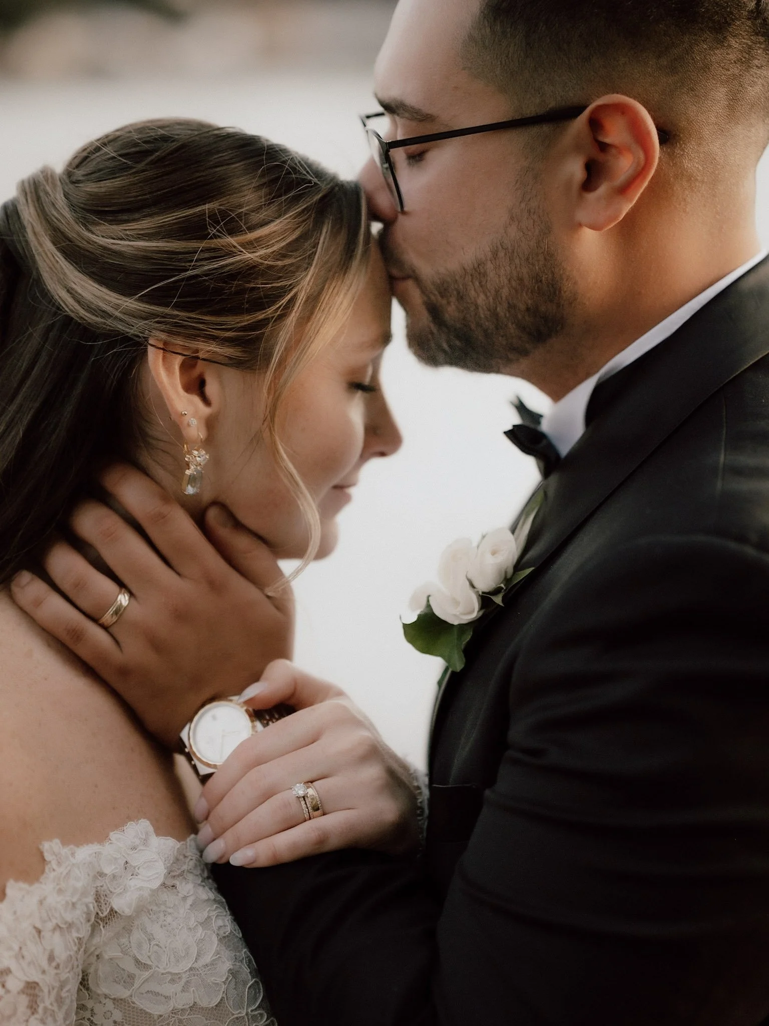 A bride and groom sharing a tender moment with foreheads touching and eyes closed, dressed in wedding attire, with the bride wearing lace dress and jewelry, and the groom in a black tuxedo with a white boutonniere.