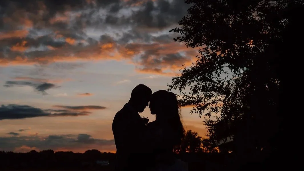 Silhouette of a couple with foreheads touching, standing outdoors at sunset with colorful clouded sky and trees in the background.