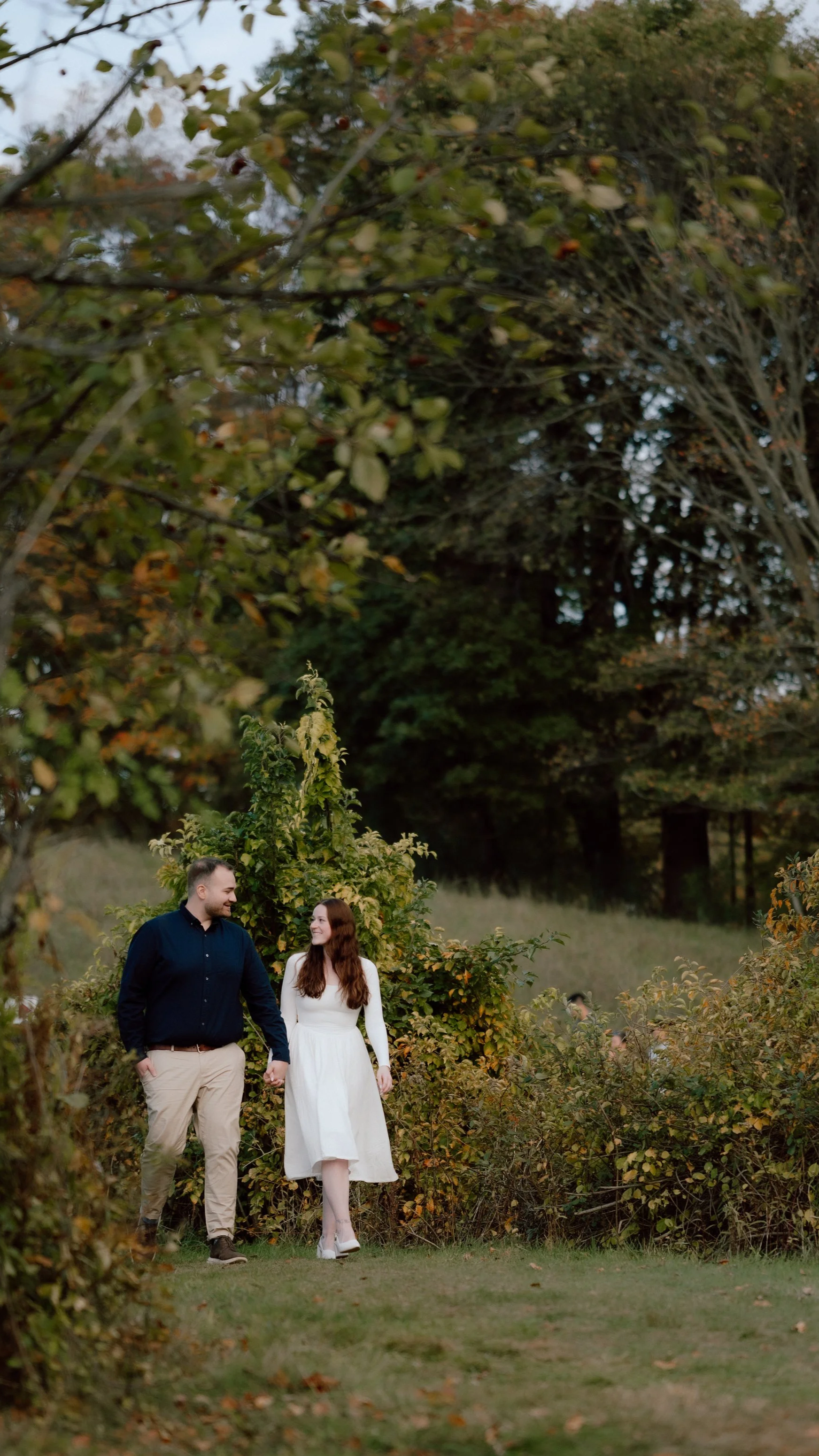 A couple walks hand-in-hand through a lush garden path at Bancroft's Castle in Groton, MA, surrounded by dense greenery and trees with autumn leaves, smiling at each other.