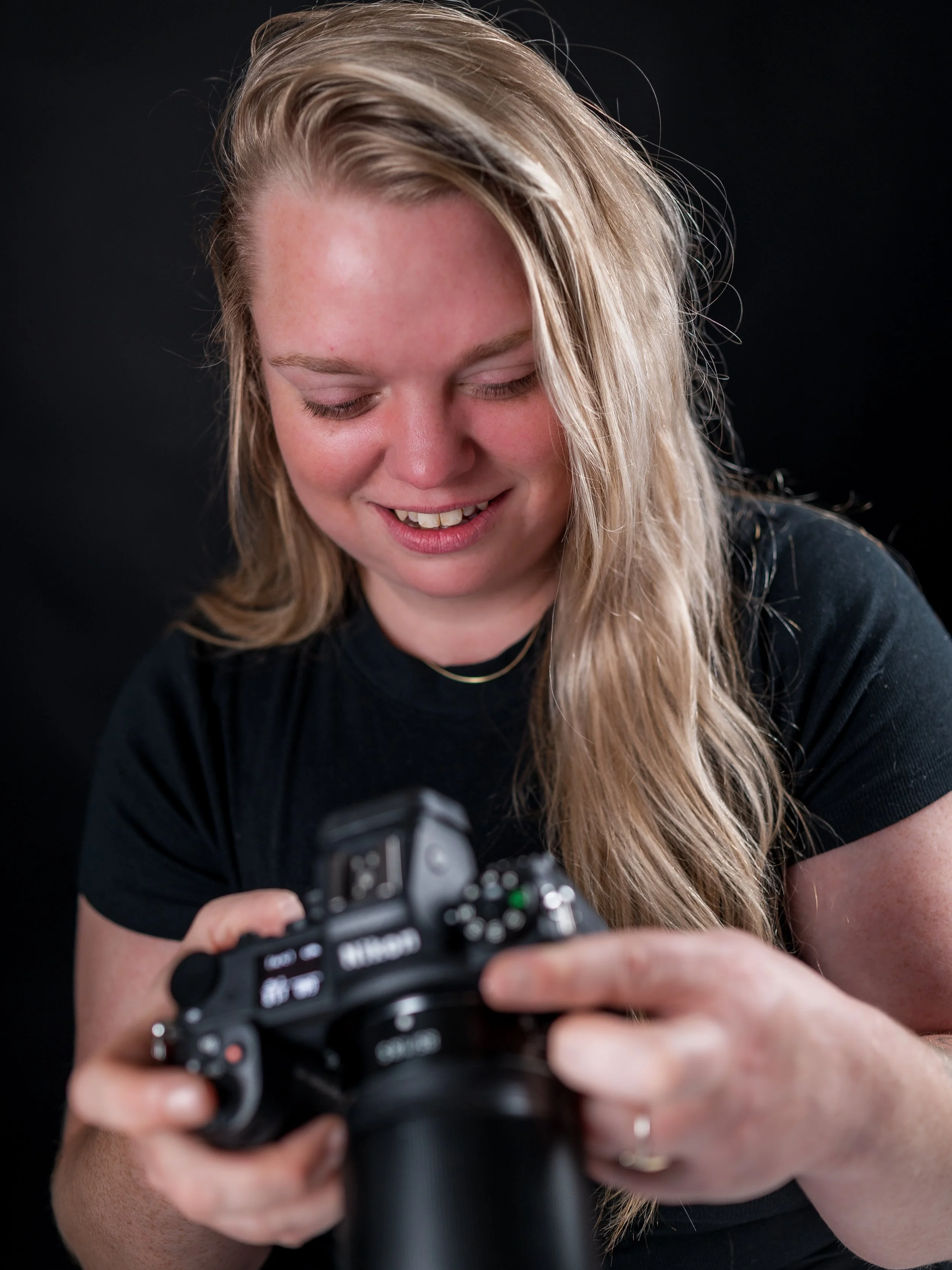 A woman with long blonde hair smiling while looking down at a camera she is holding.