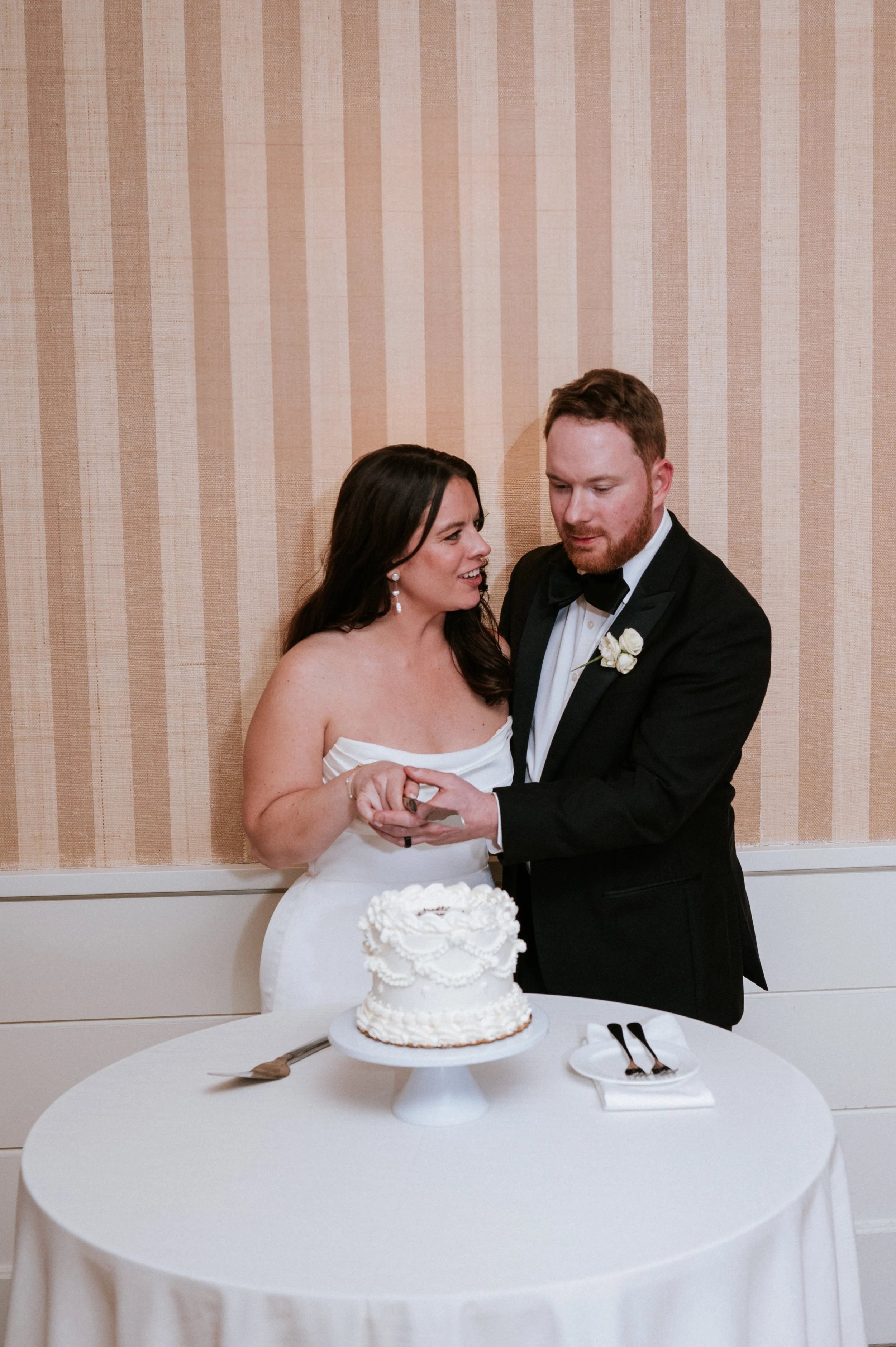 A newlywed couple in formal attire cutting a wedding cake at their reception.