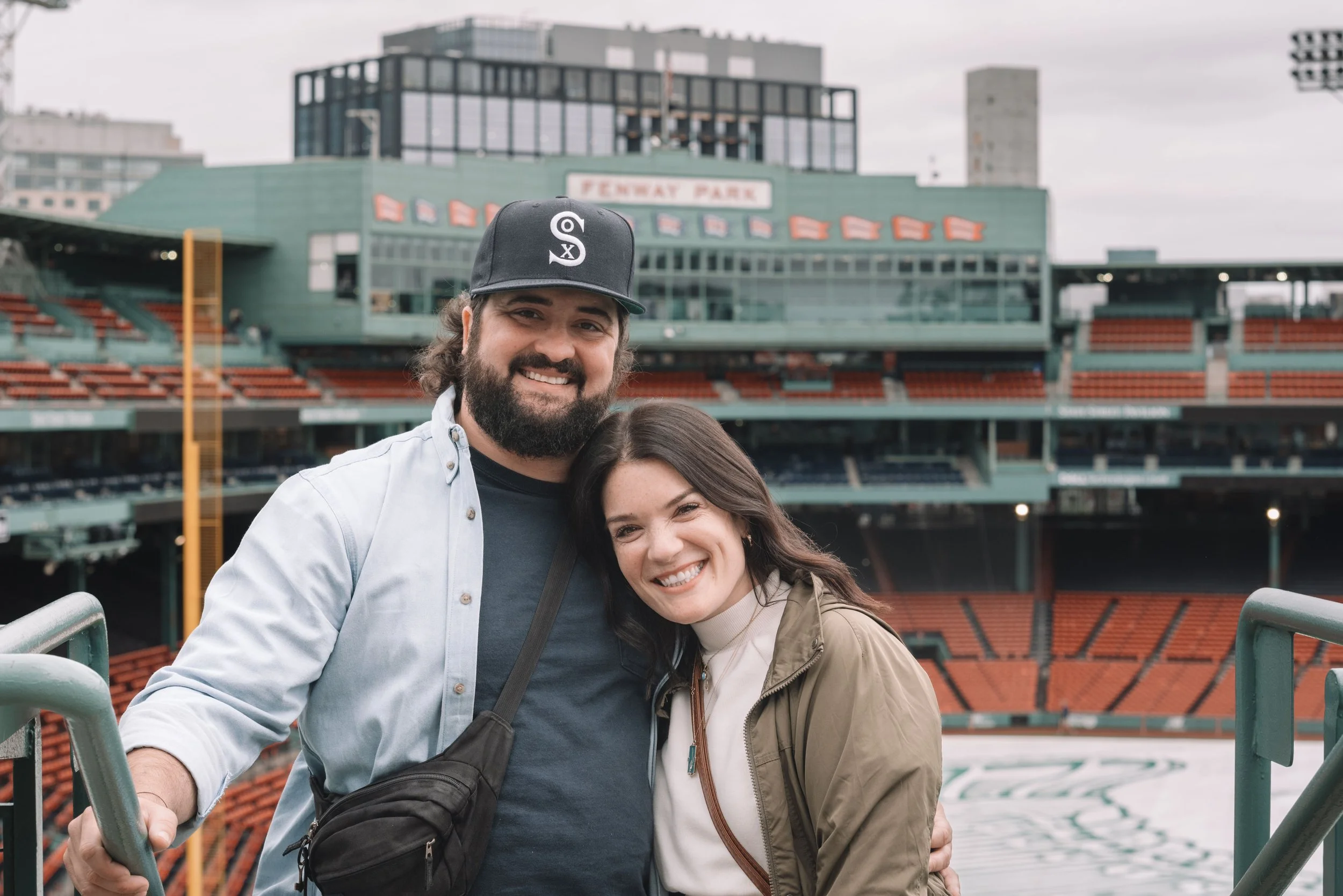 A smiling man and woman standing in a stadium, with the field and seating area visible behind them. The man has a beard, wearing a baseball cap and a light shirt. The woman has long dark hair, wearing a light-colored turtleneck and a jacket.