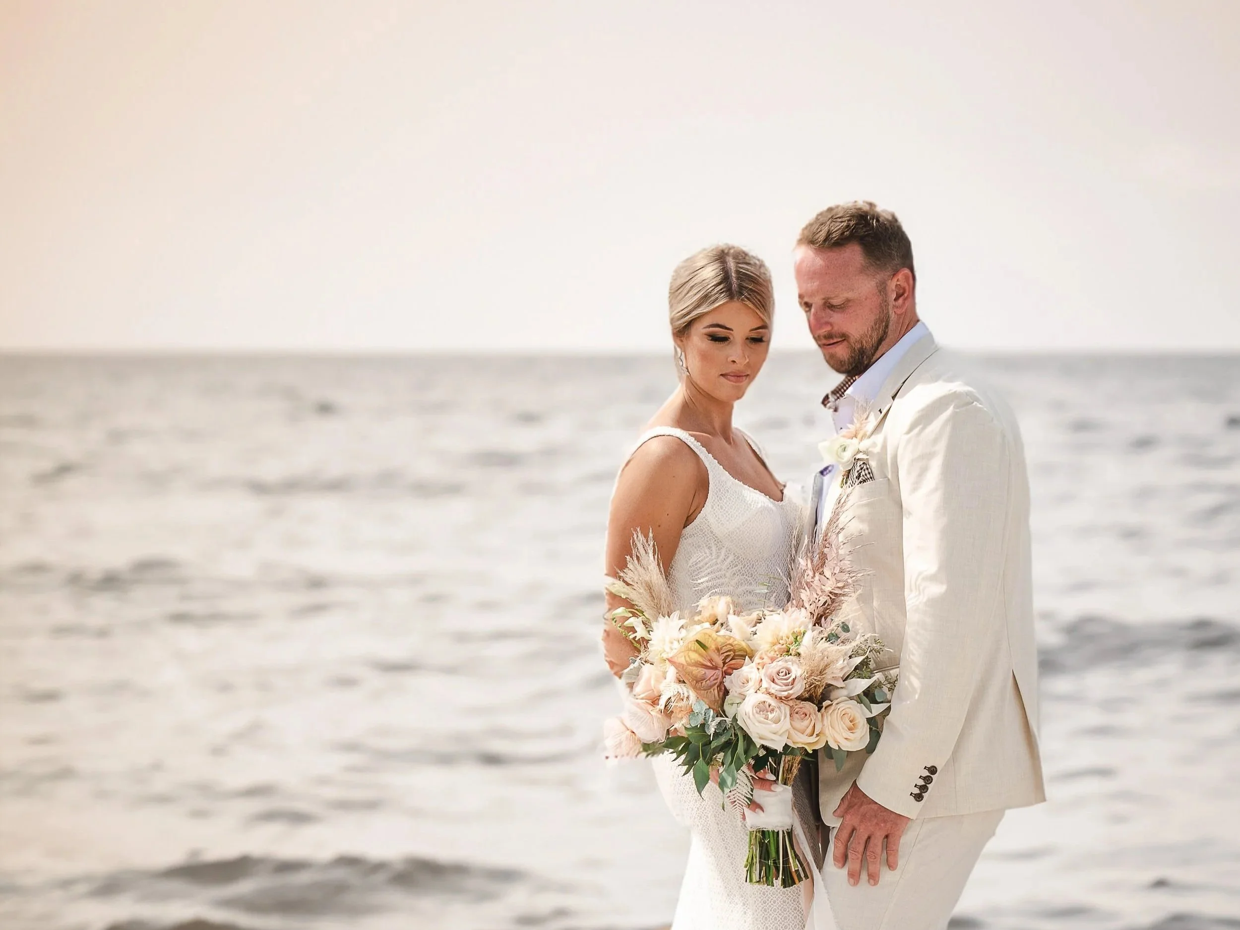 A  Newport, Rhode Island bride and groom in wedding attire standing close together on a beach, holding a bouquet of flowers, with the ocean in the background.