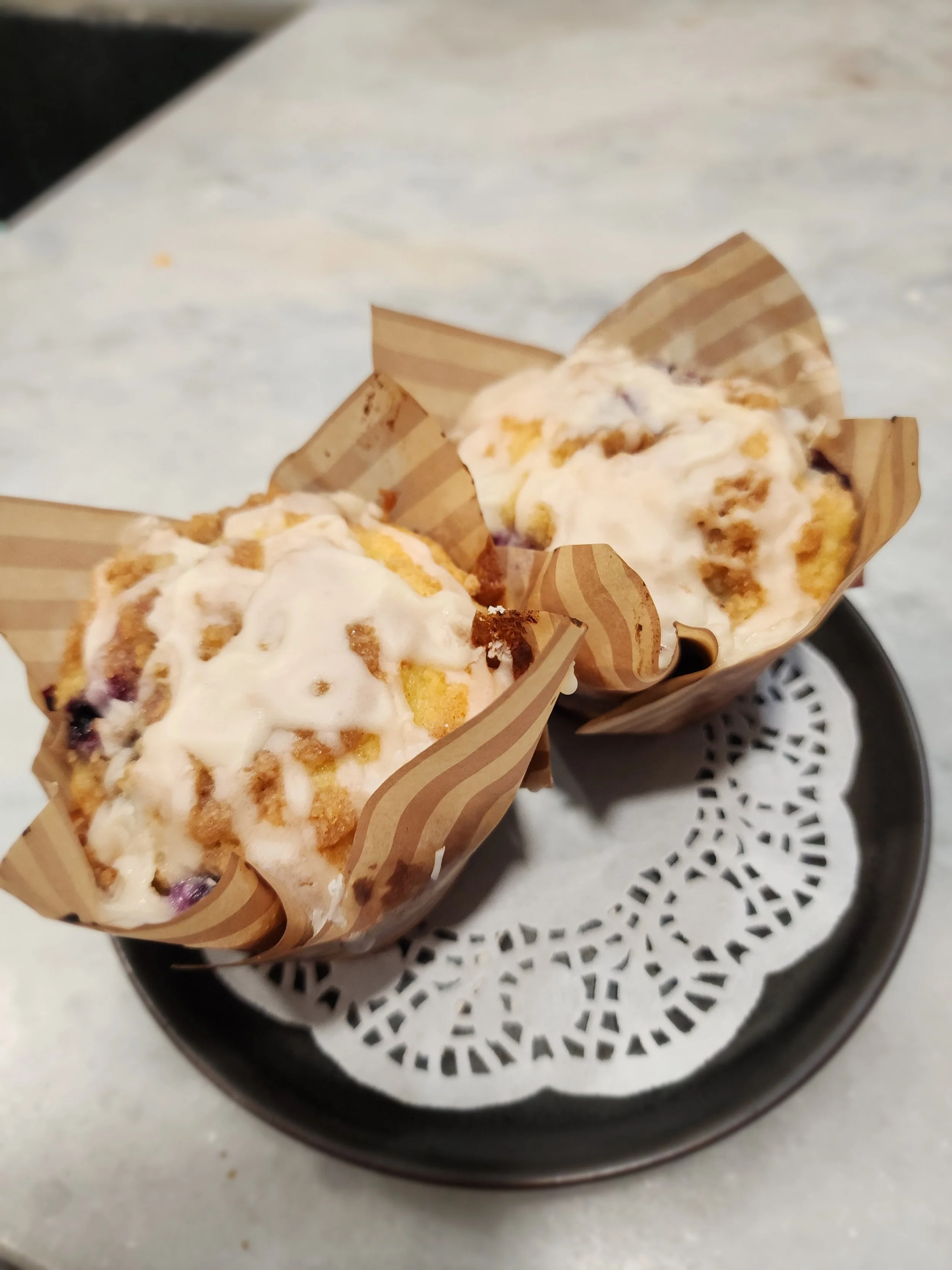 Two muffins with white icing and crumb topping in paper wrappers on a black plate with a paper doily.