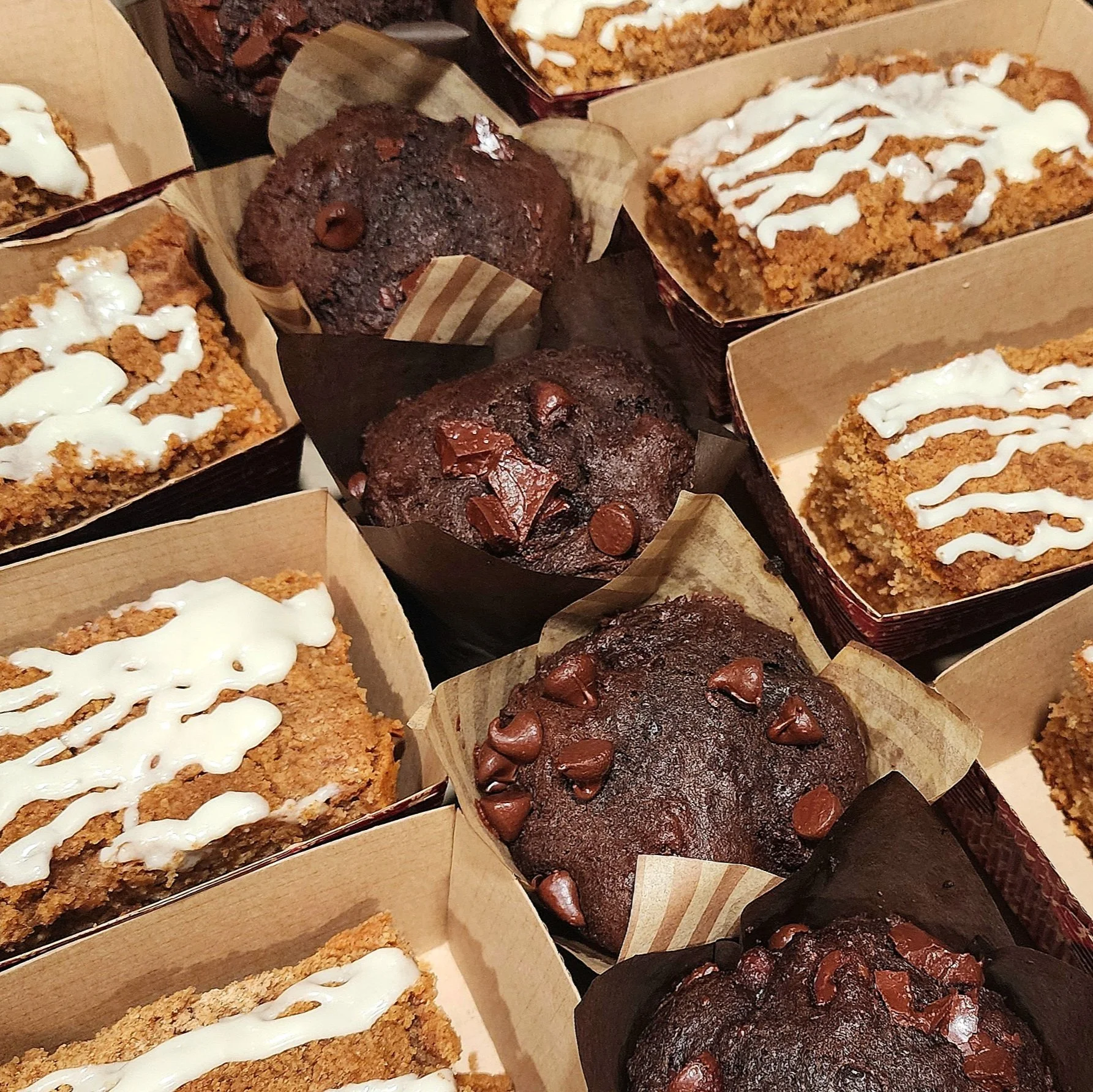 Assorted brownies and chocolate muffins in paper trays, some drizzled with white icing and others with chocolate chips.