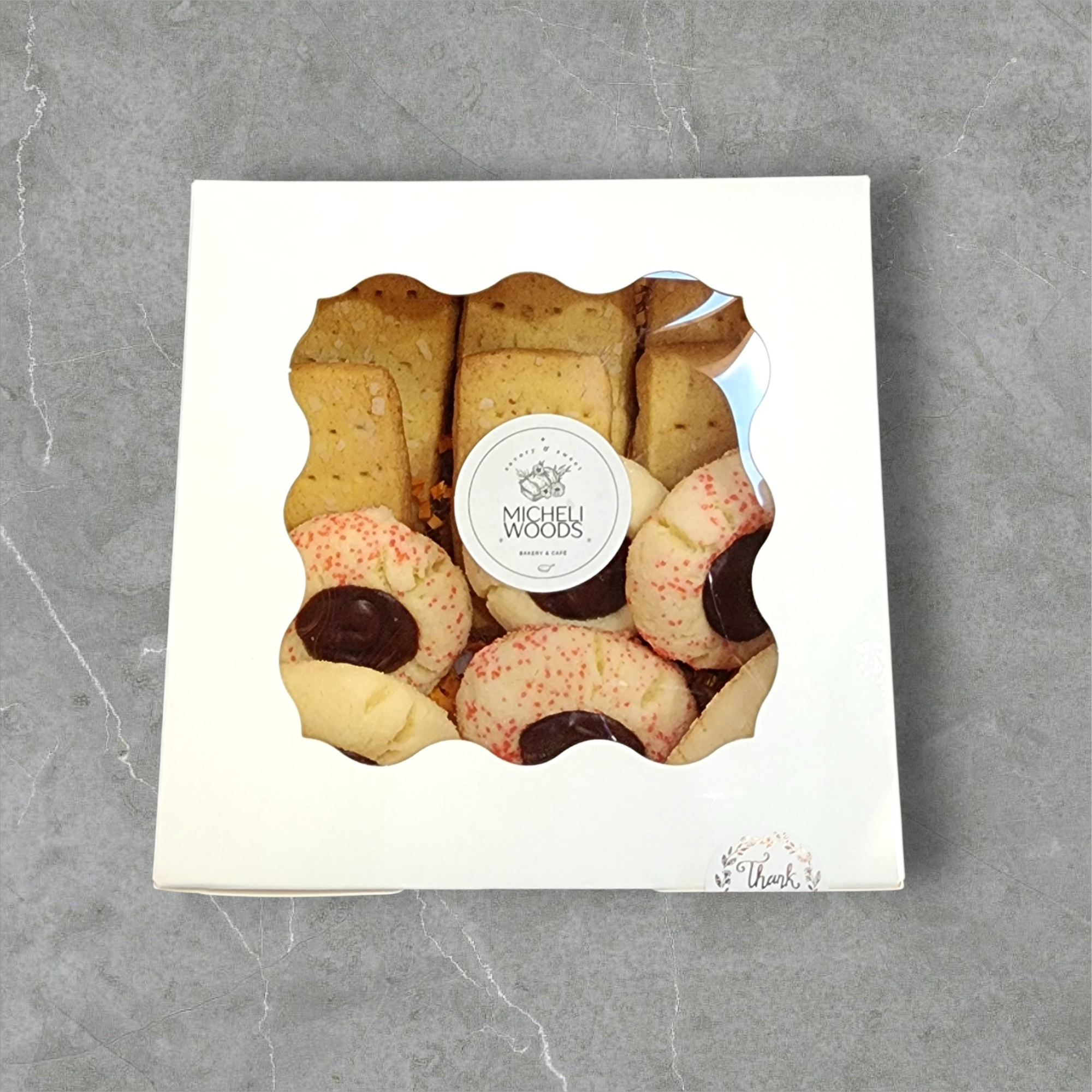 A white box of cookies and pastries from Michel Woods bakery, seen through a decorative window in the box, placed on a gray stone surface.