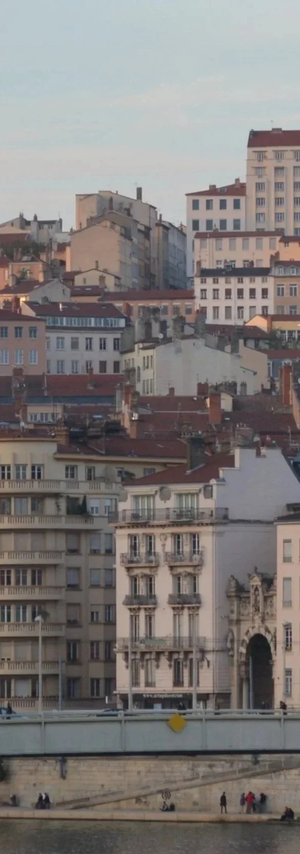 Une vue urbaine avec plusieurs bâtiments résidentiels en hauteur, certains avec des toits en pente rouges, et un pont au premier plan au-dessus d'une rivière, sous un ciel clair.