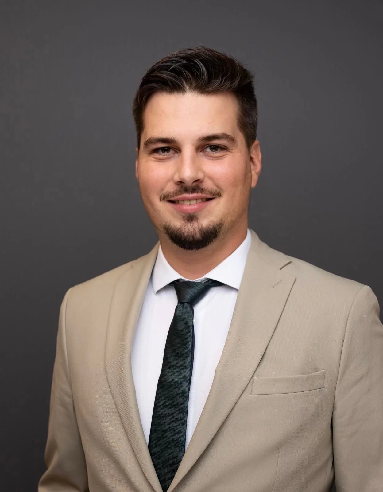 Portrait d'un homme d'affaires en costume beige, chemise blanche et cravate noire, souriant, contre un fond gris