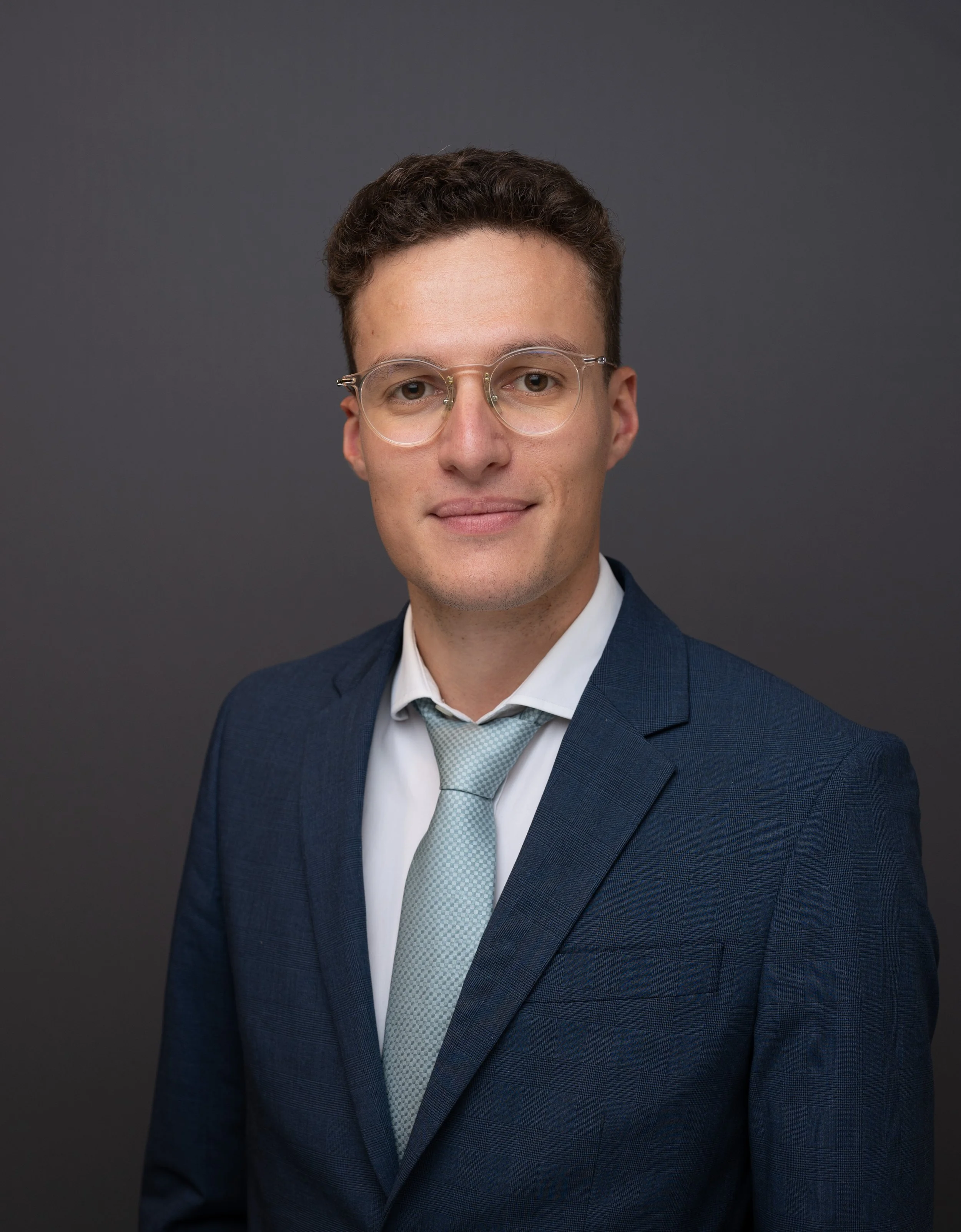 Portrait d'un jeune homme en costume bleu, chemise blanche, cravate gris clair, portant des lunettes, contre un fond gris foncé.