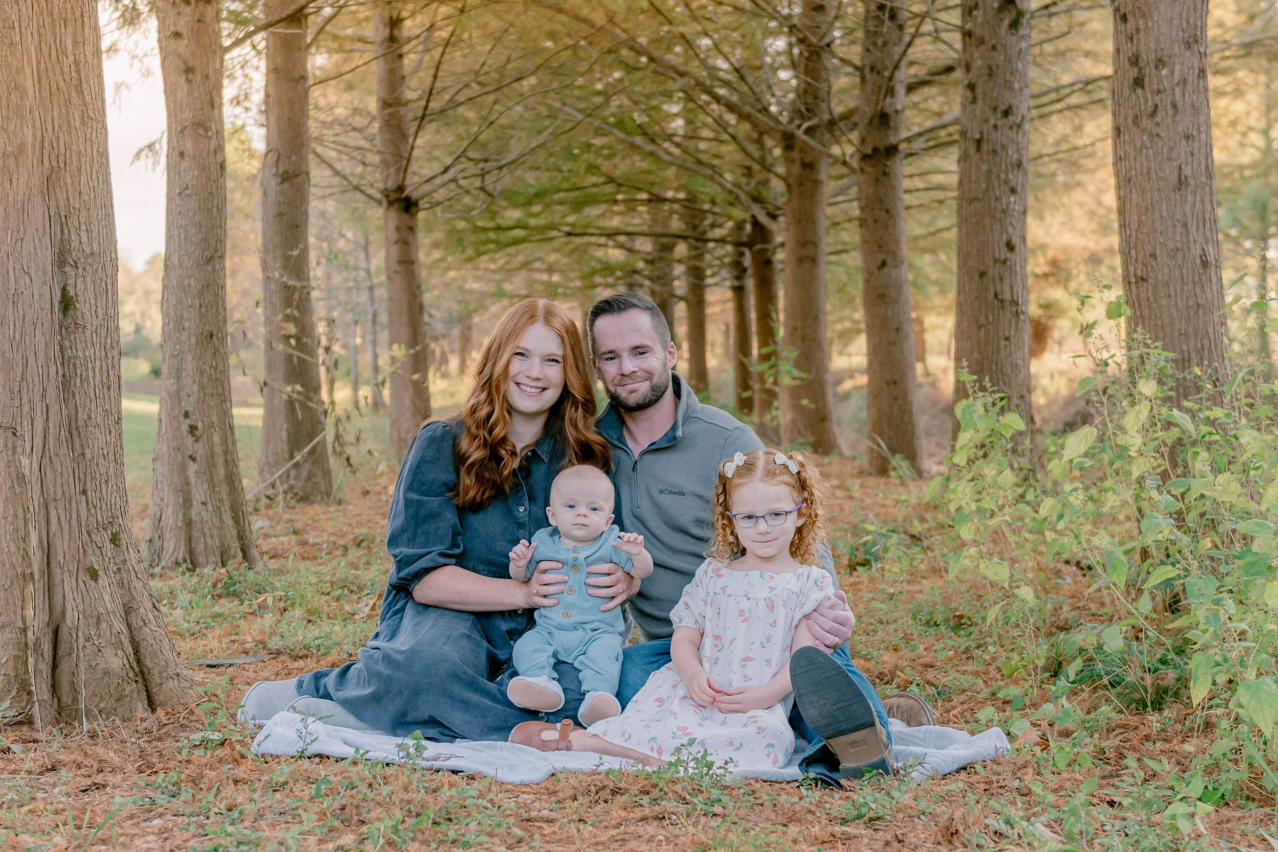 A family of four sitting on a blanket in a wooded area during fall. The mother has red hair, and the father has dark hair. The older girl has curly red hair and glasses, and the baby has light hair.
