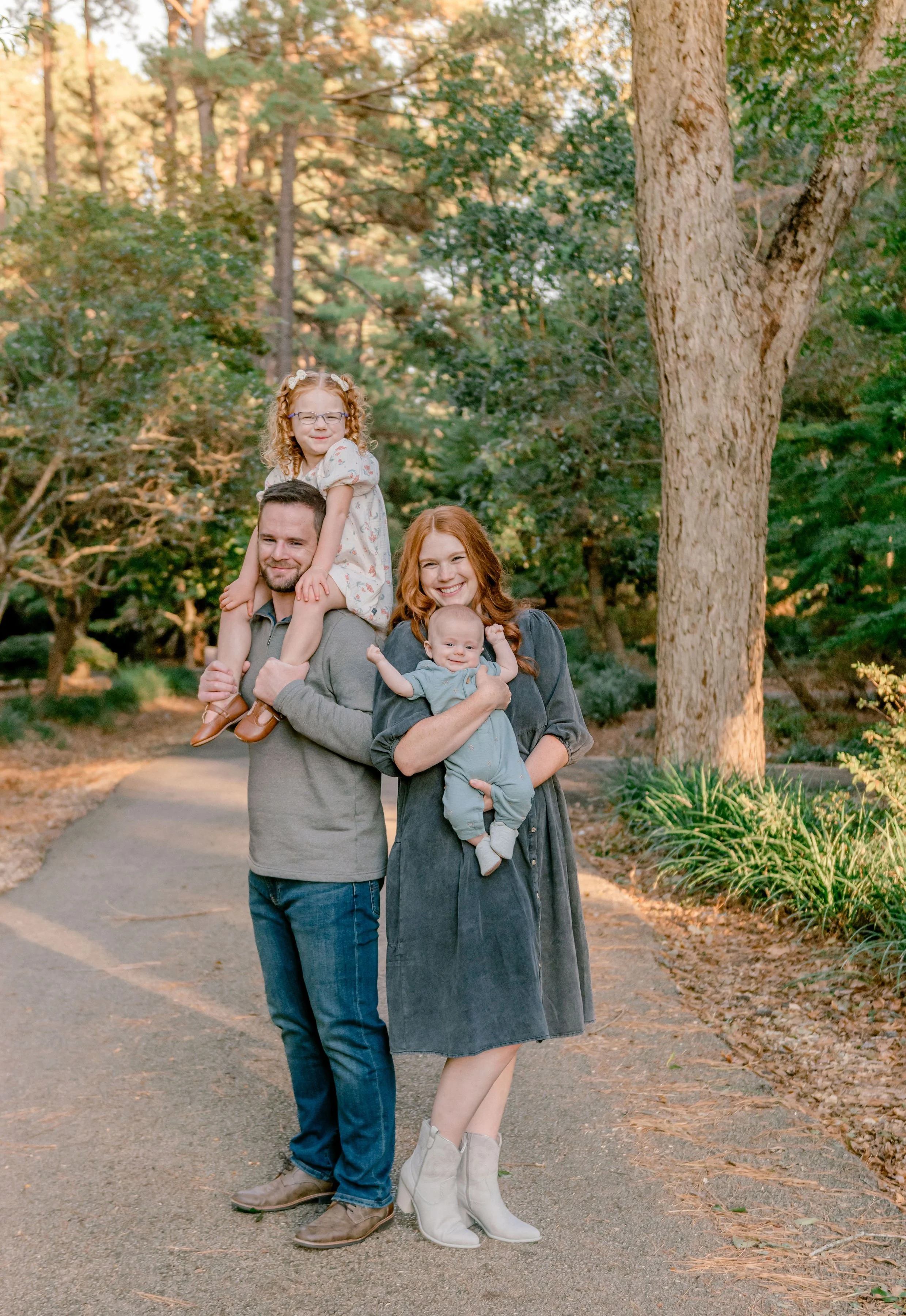 A family of four standing on a pathway in a park with trees and greenery, smiling at the camera. The father is carrying a young girl on his shoulders, and the mother is holding a baby.