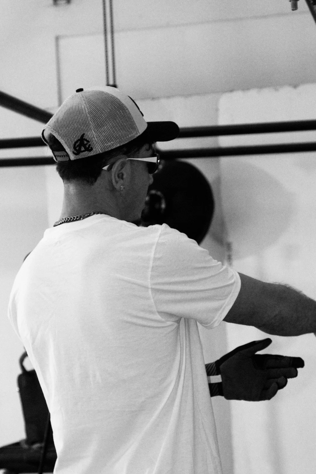 A person wearing a baseball cap, sunglasses, a white t-shirt, and gloves, standing in a gym or workout space near a weightlifting bar.