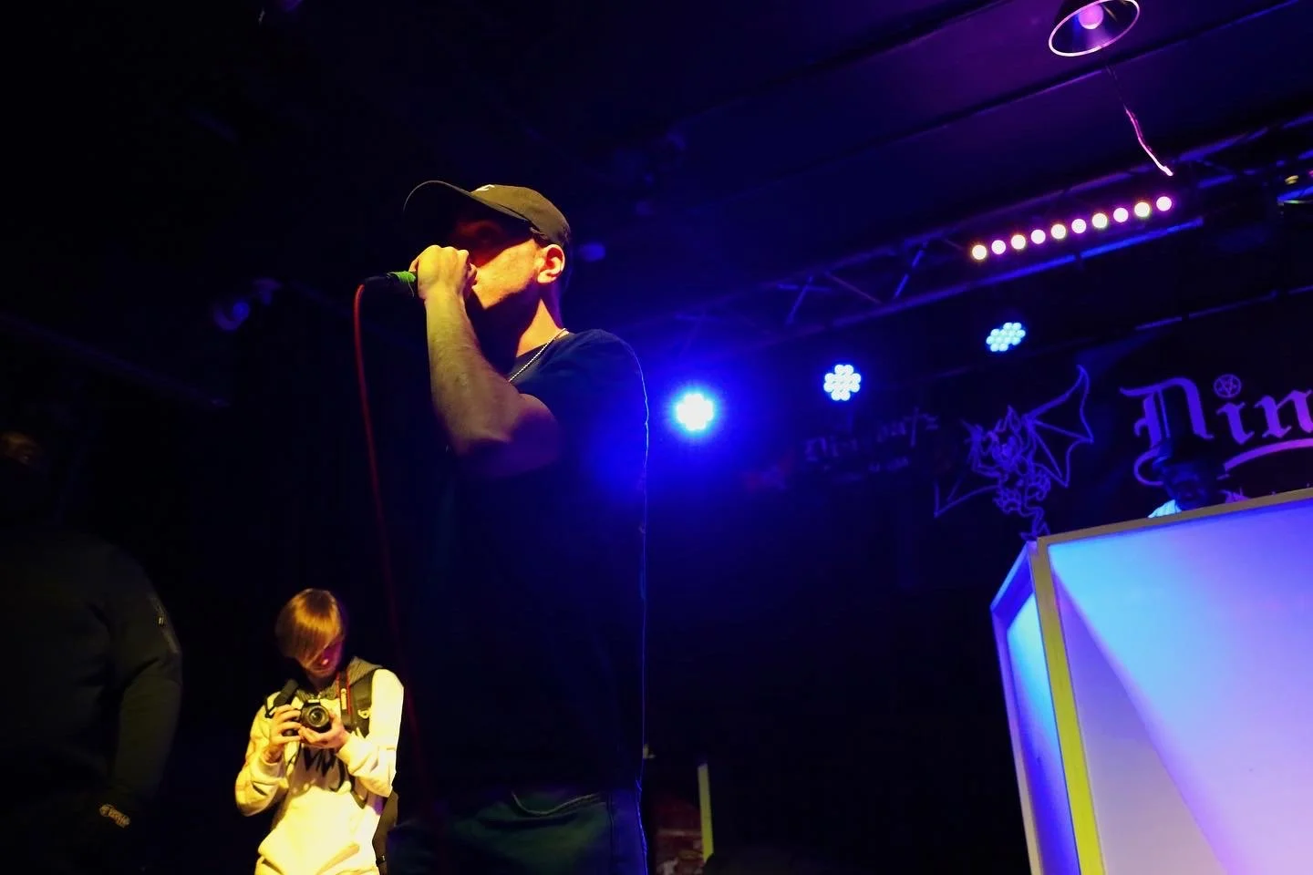 Person wearing a cap singing into a microphone on stage, with blue and purple stage lights, and a photographer taking pictures.