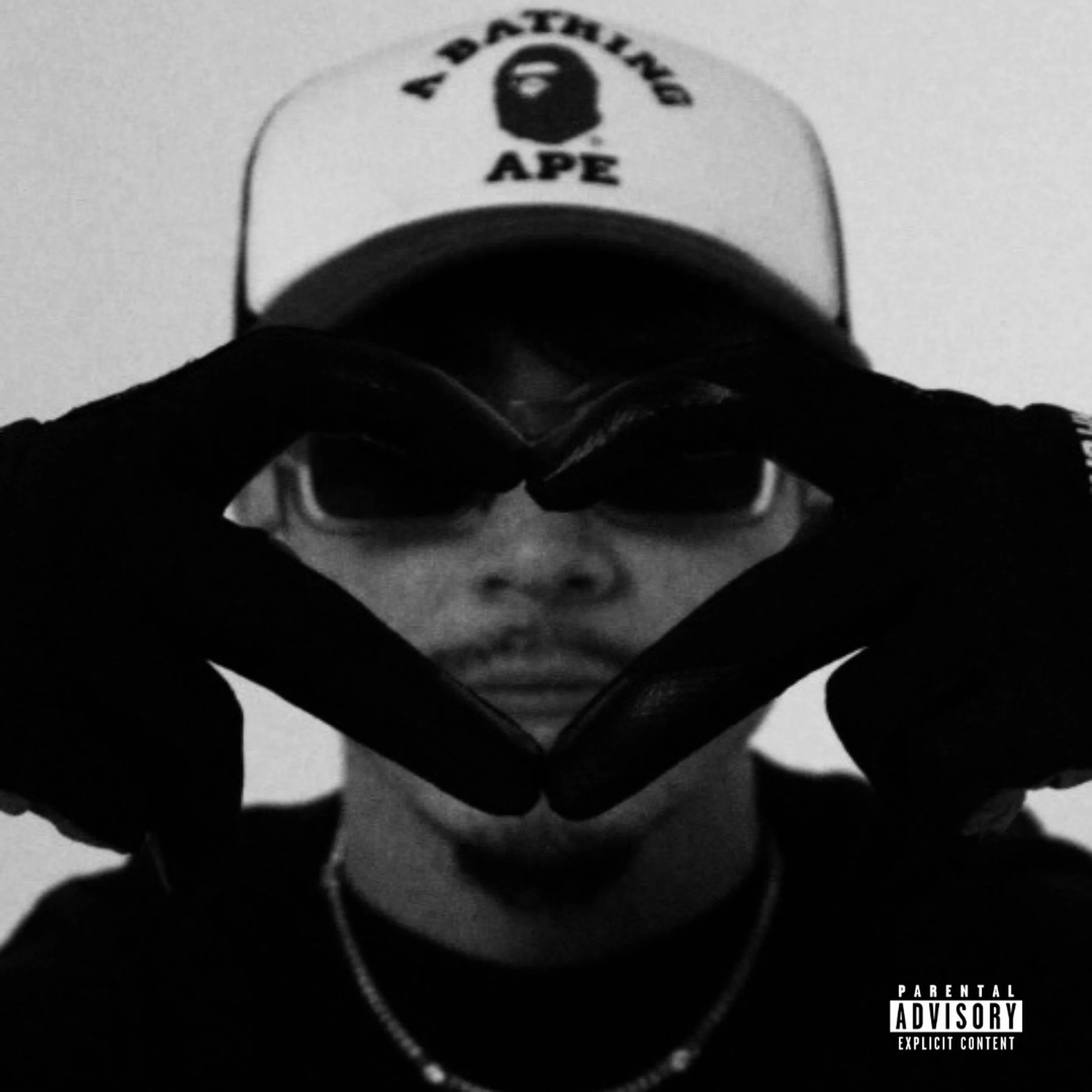 Black and white photo of a person wearing Hatsune Miku glasses, a beige cap with 'A BATHING APE' logo, and gloves forming a heart shape around their face.