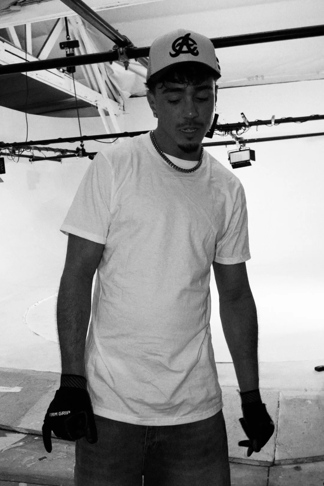 A young man wearing a baseball cap, white t-shirt, and gloves is standing in a photography or film studio with lighting equipment overhead.
