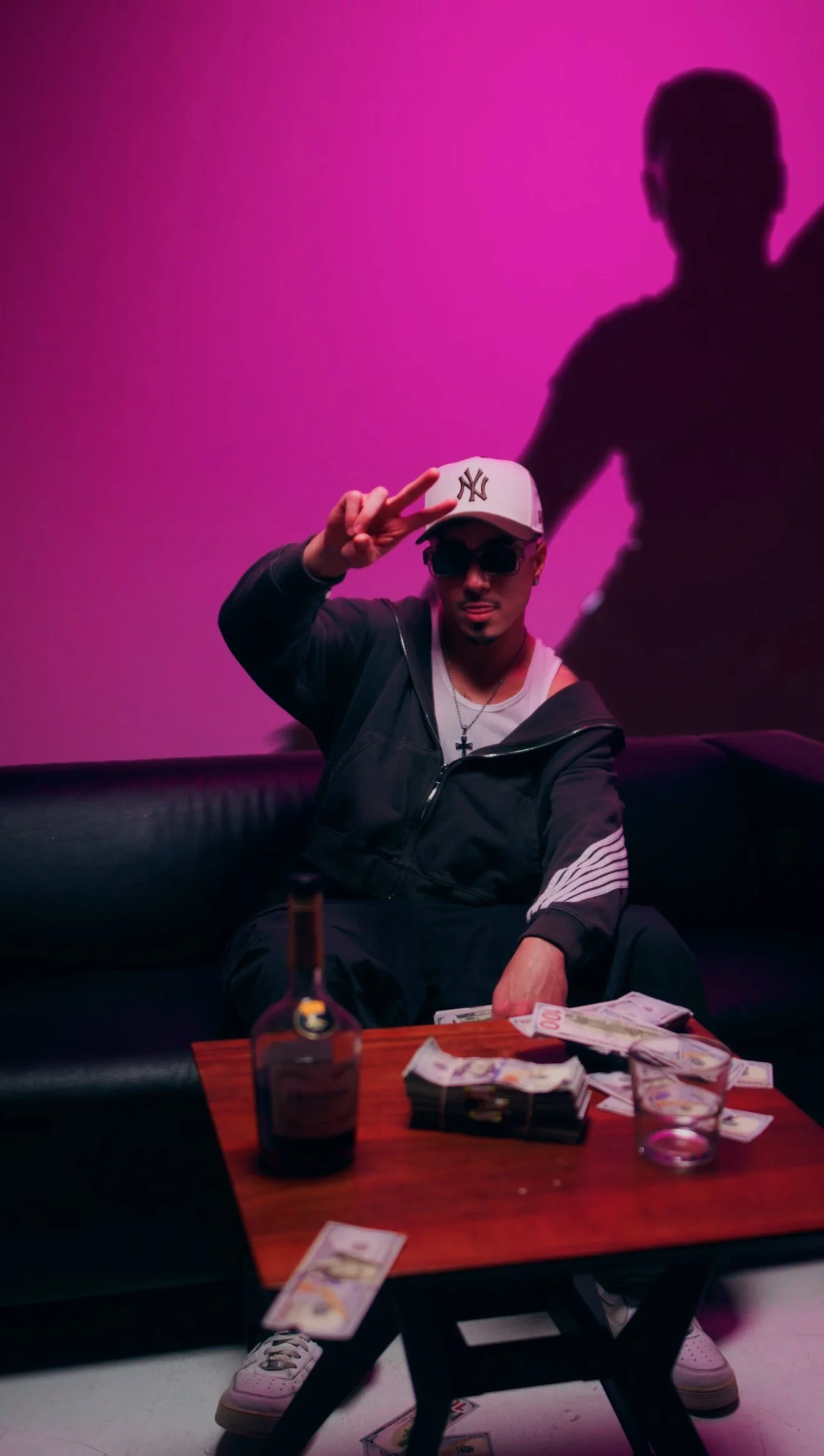 A man wearing a white NY Yankees cap, sunglasses, and a black jacket making a peace sign while sitting on a black couch with a table of cash, a bottle of alcohol, and a glass in front of him. There is a silhouette of another person on the pink background behind him.