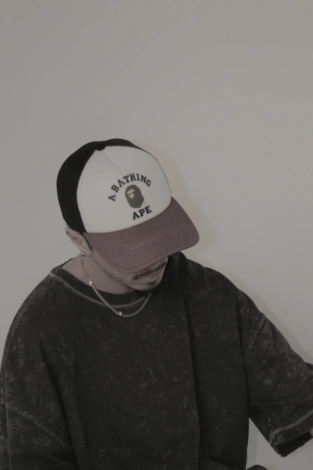 A person wearing a black and white baseball cap with a logo that says "A Bathing Ape" with a face illustration, a black patterned shirt, a silver chain necklace, and standing against a plain light-colored wall.