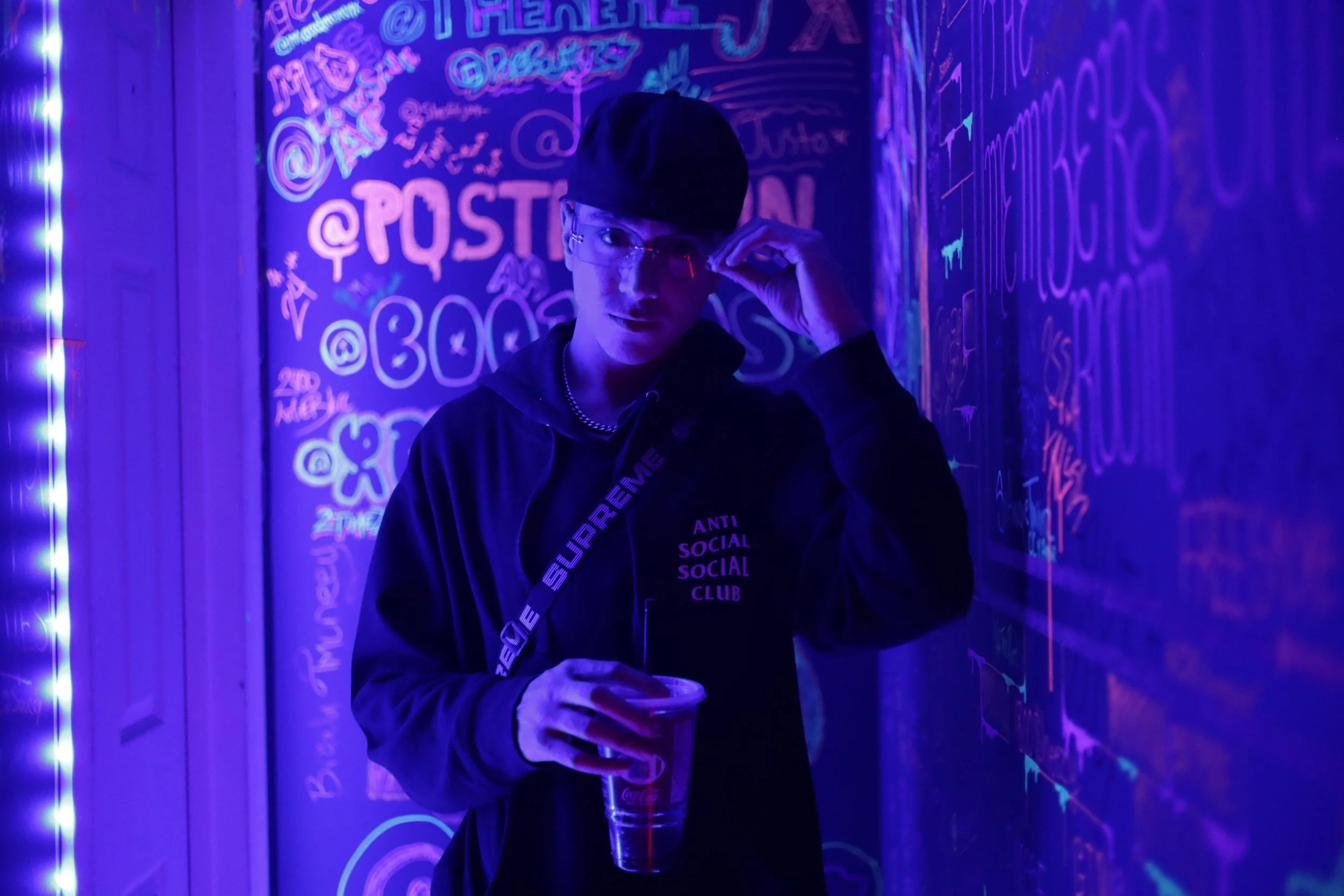 A person wearing glasses and a black cap, dressed in a black hoodie, stands in front of a wall covered in colorful neon graffiti and writings. The person is holding a drink in one hand and adjusting their glasses with the other. The scene is illuminated by purple and blue neon lights.