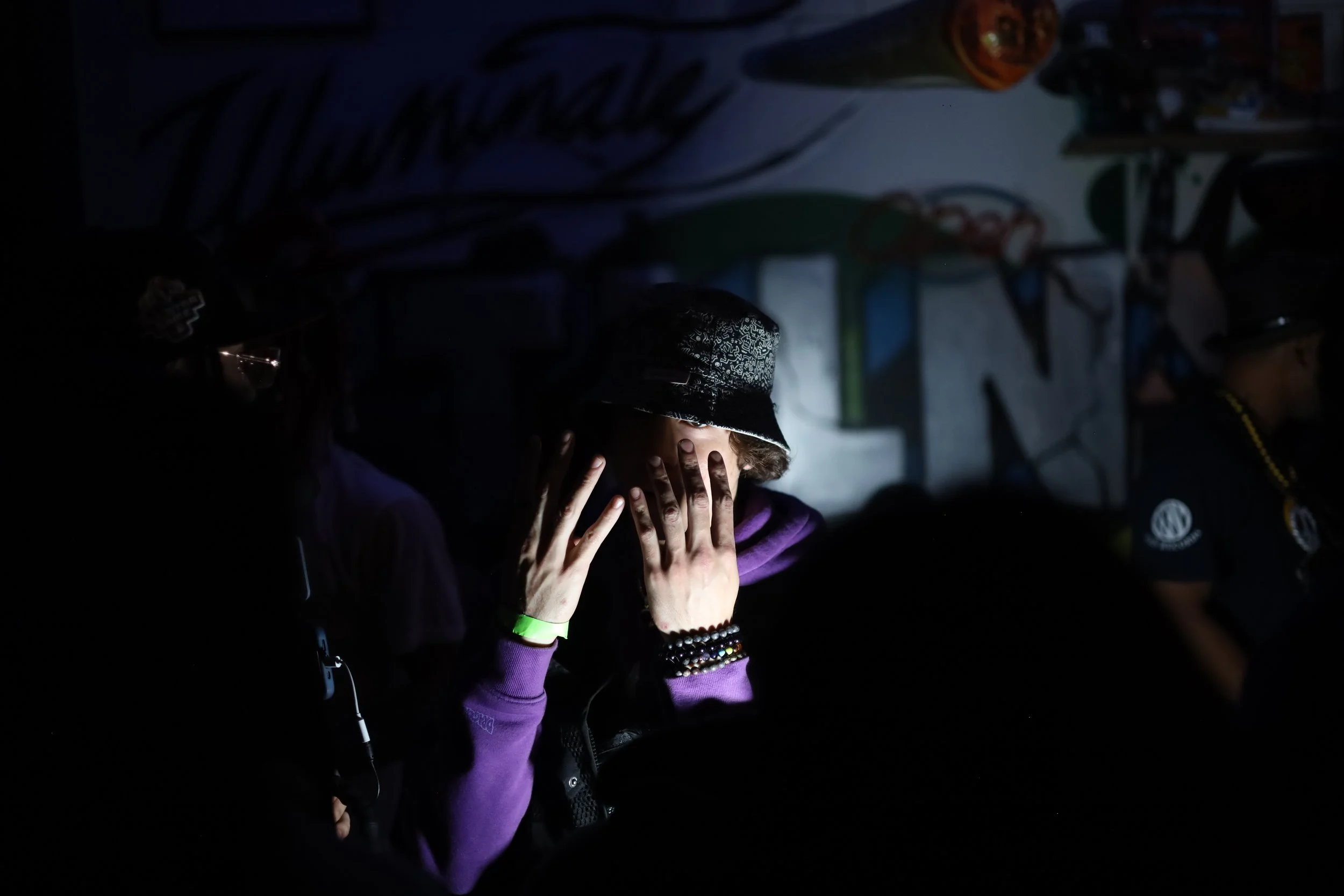 Person wearing a bucket hat covering their face with both hands, in a dark room with graffiti on the wall behind.