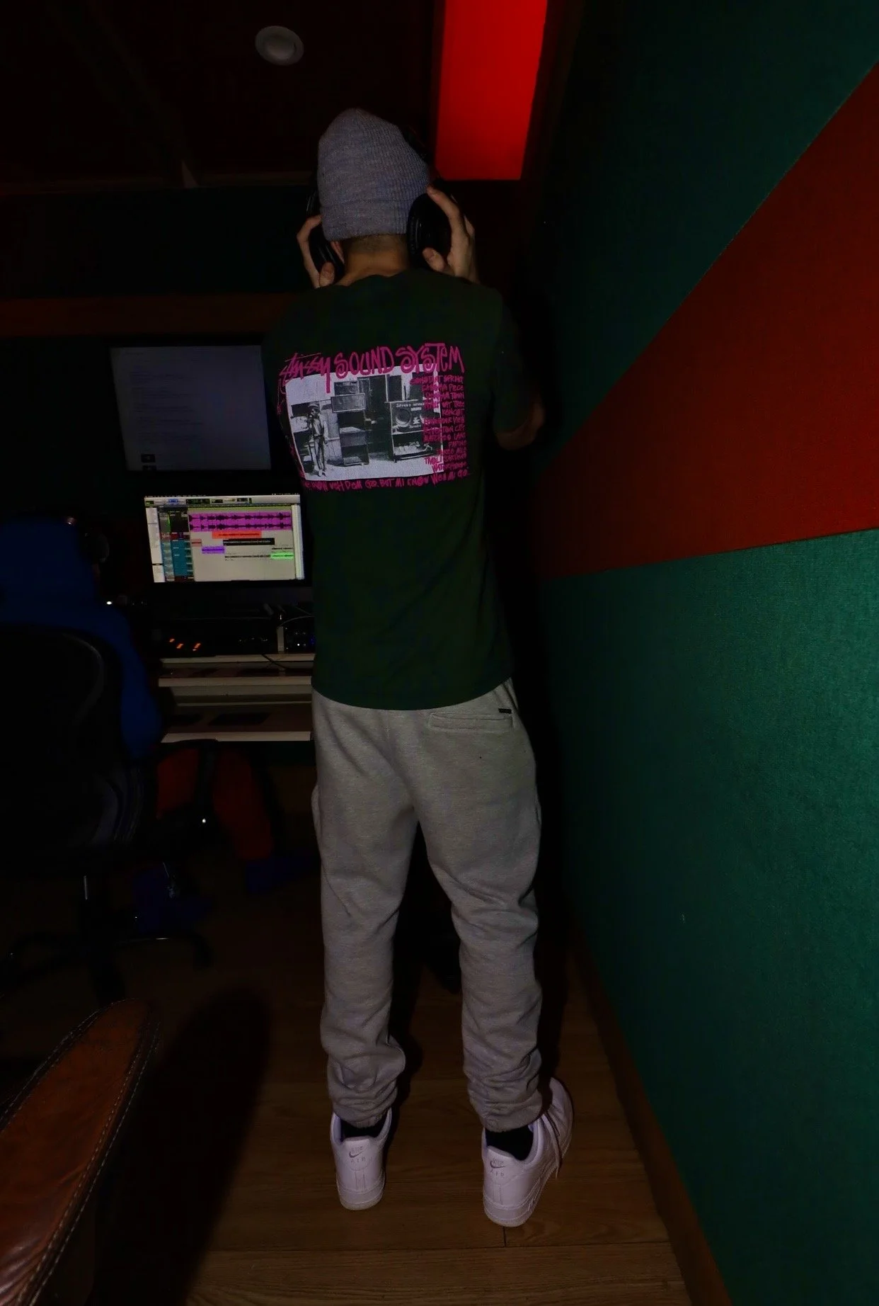 A person standing in a music studio, wearing a gray beanie, black T-shirt with pink and black graphic on the back, gray sweatpants, and white sneakers, with large headphones on.