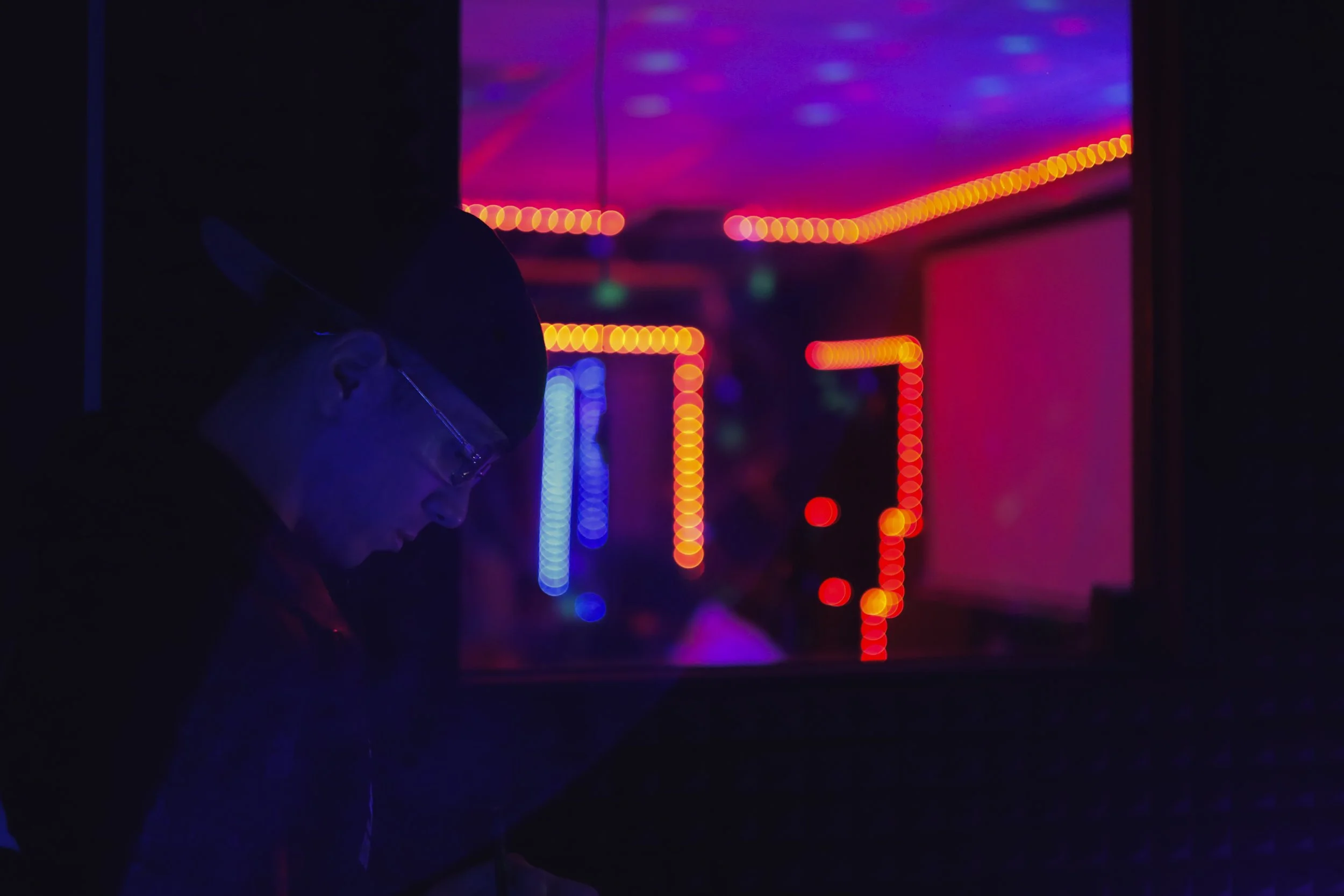 Person wearing glasses and cap in a dark room with neon pink, purple, red, and blue LED strip lights.