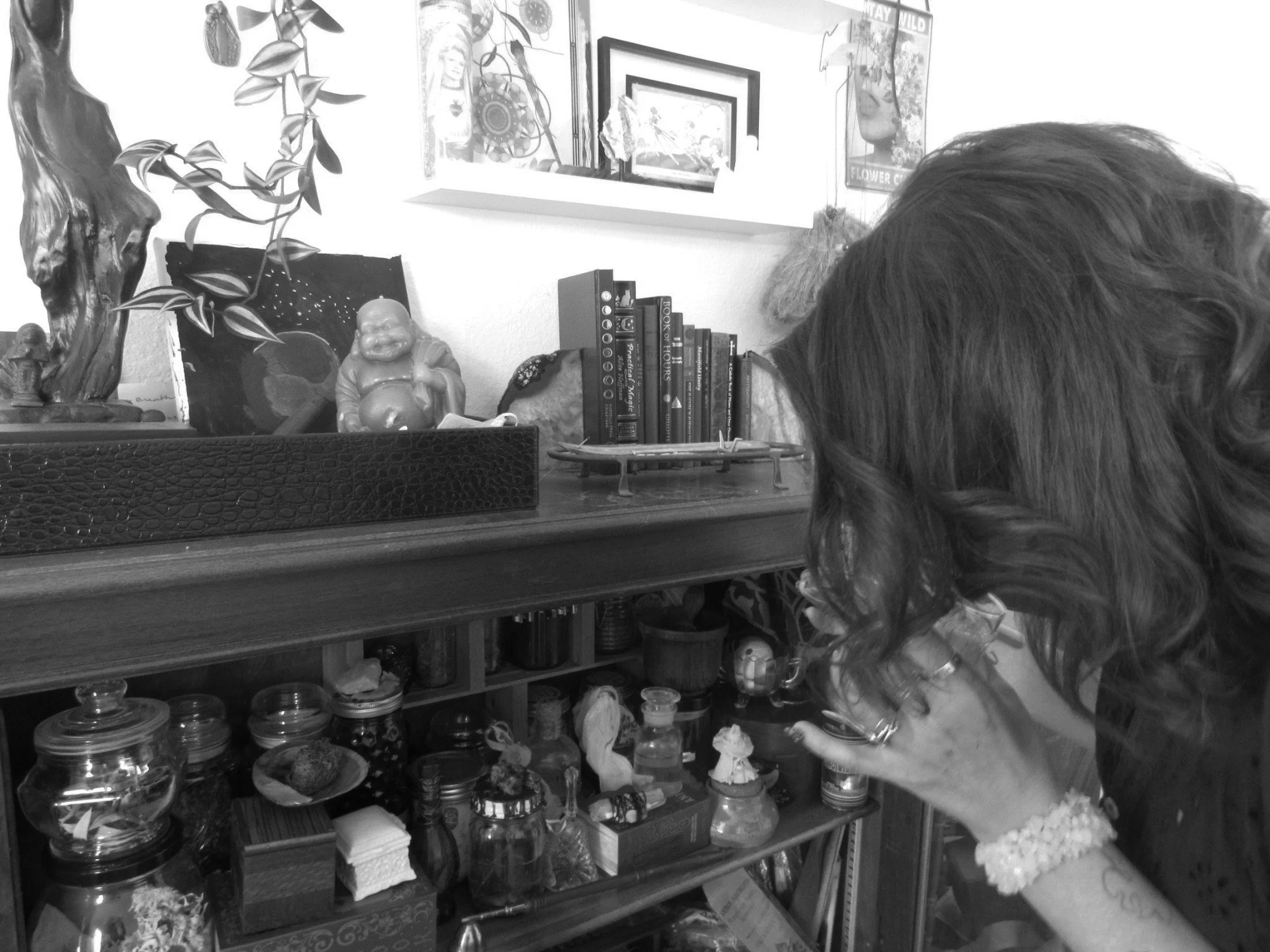 A woman with wavy hair and a bracelet on her wrist looking at a display of small jars and collectibles on a wooden shelf in a room decorated with framed pictures, plants, and various decorations.