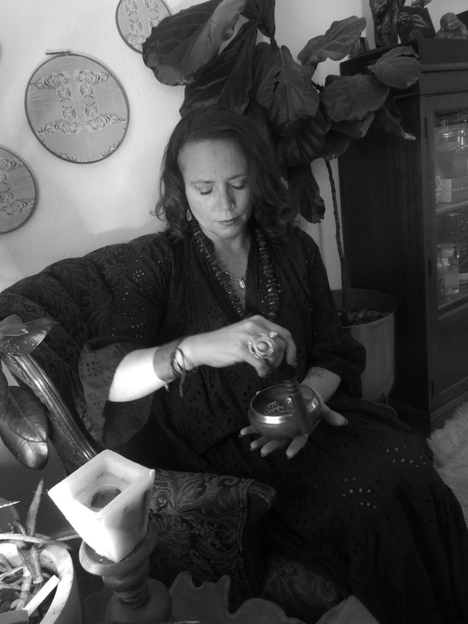 A woman with dark wavy hair, wearing a nose ring, jewelry, and a dark blouse, is sitting on a patterned armchair, stirring a small bowl with a spoon in her hand. She appears focused on her task. Behind her, there are decorative wall hangings with tiger designs, a large plant with broad leaves, and a cabinet with glass doors. In the foreground, there is a candle holder with a square candle and a potted plant.