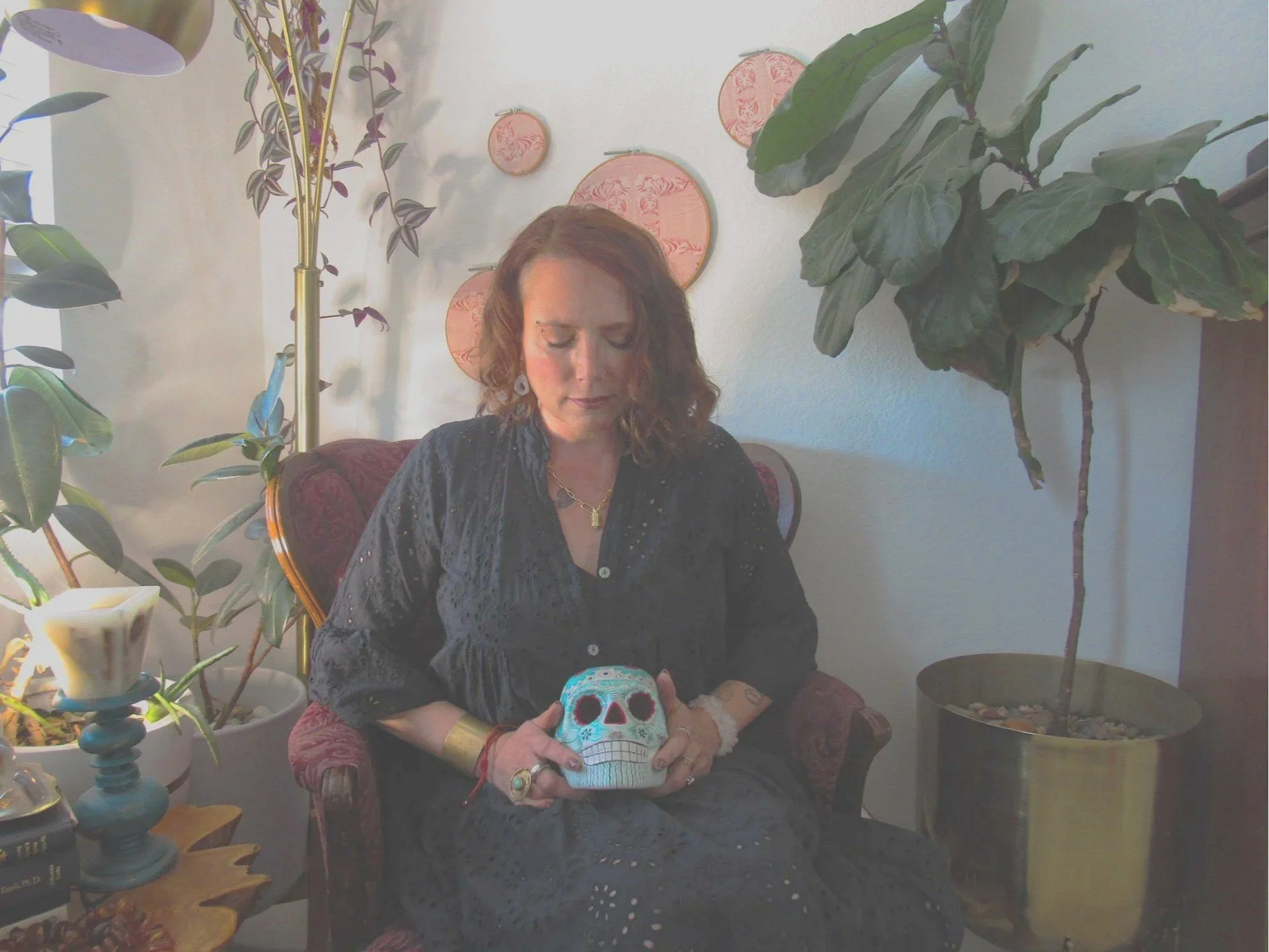 A woman with reddish-brown hair sitting in a red and gold armchair, holding a decorative skull, surrounded by houseplants and wall art.