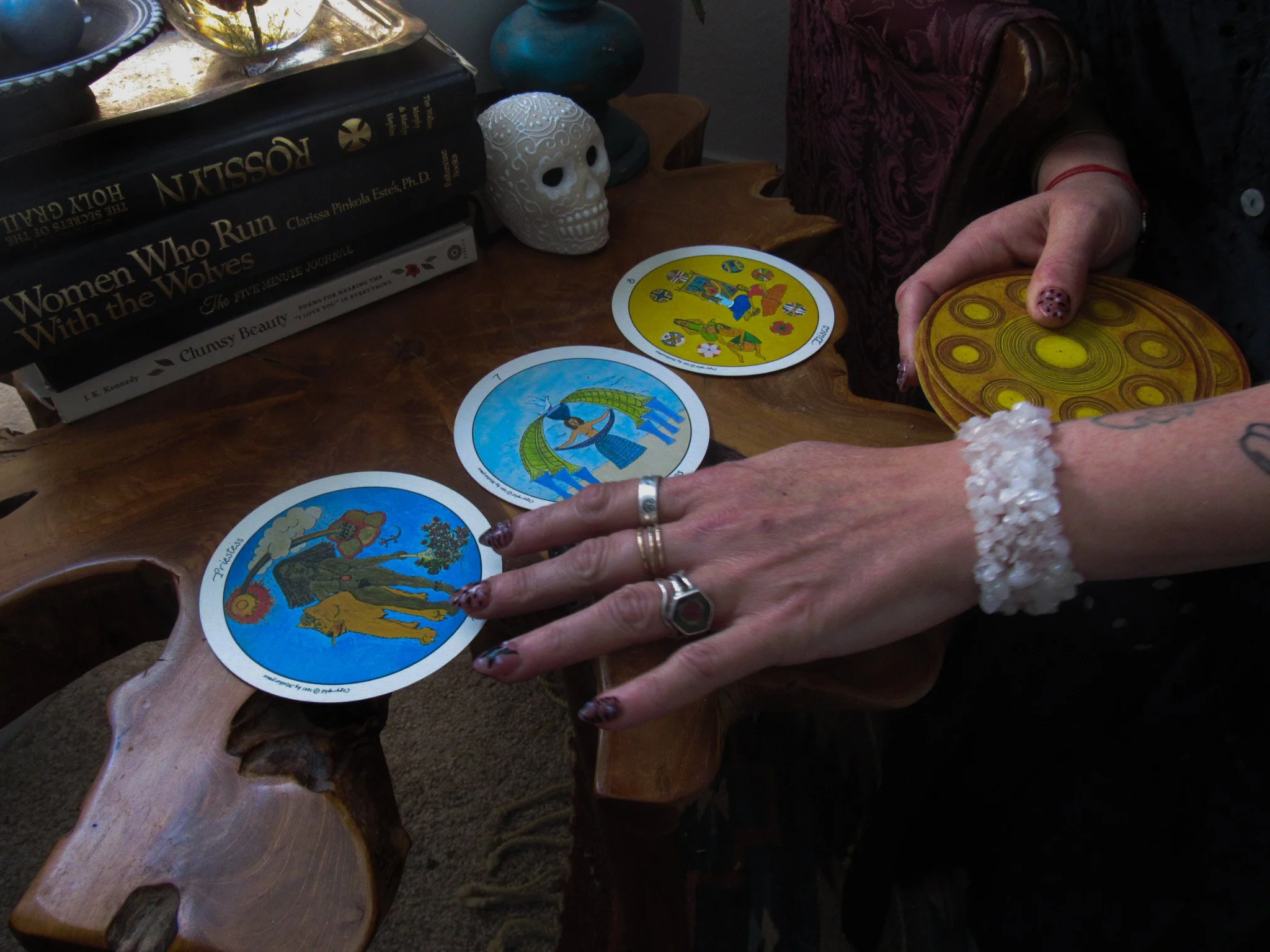 A person is holding Tarot cards spread on a wooden table, with additional cards in their other hand. The table also has books stacked on one side and a decorative skull on the surface.
